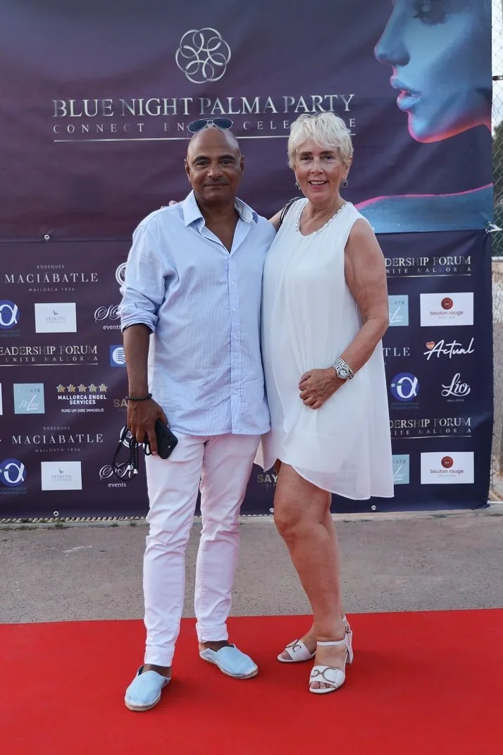Two people standing on a red carpet in front of a backdrop for the Blue Night Palma Party. The man is wearing a light blue shirt, white pants, and white shoes, and is holding sunglasses and a phone. The woman is dressed in a white sleeveless dress and white heels, with short blonde hair and a watch on her left wrist.