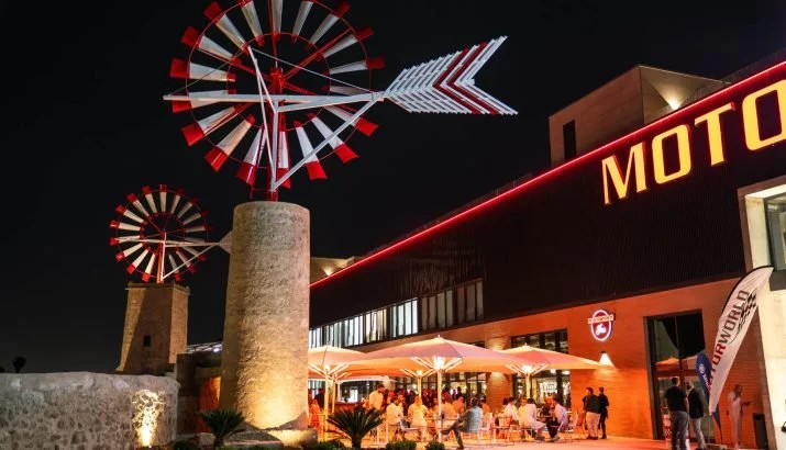 Vista nocturna de un moderno restaurante o local de ocio con estructuras de molinos de viento iluminadas sobre torres y un letrero que dice "MOTO". Hay gente cenando al aire libre bajo sombrillas.