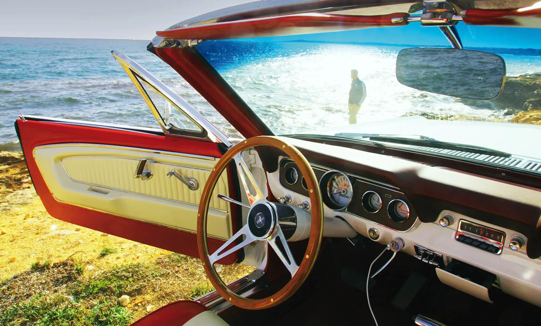 Interior of a vintage car parked near the coast with the door open, showing the steering wheel, dashboard, and part of the door panel, with the sea and a person in the background