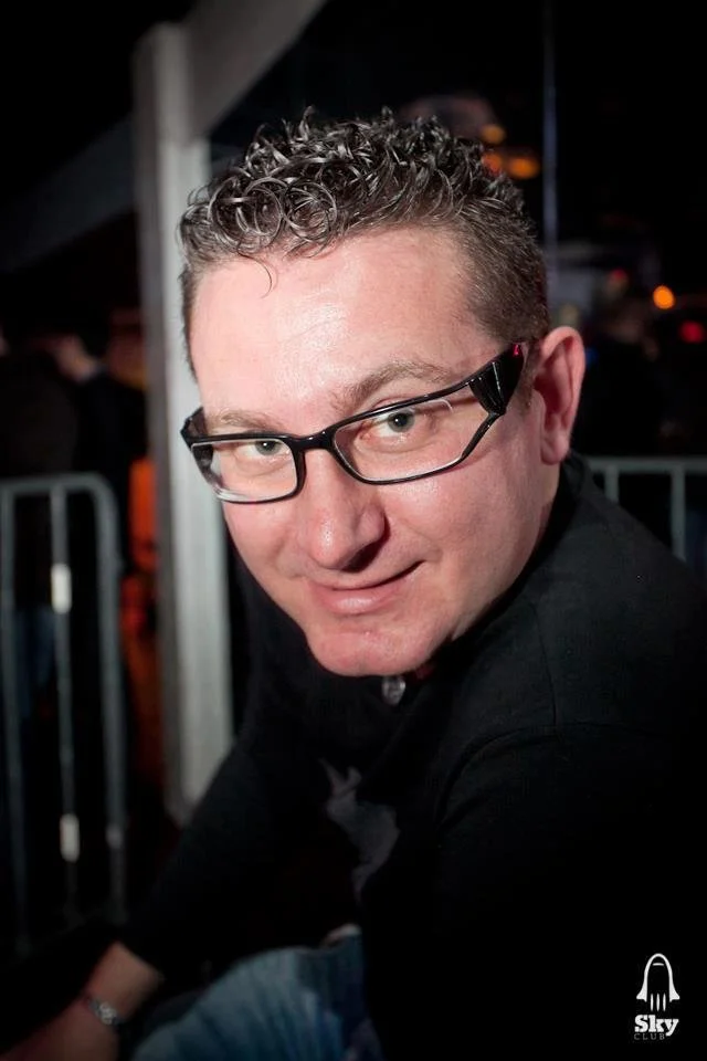 A man with curly hair and glasses smiling at the camera in a dark indoor setting.