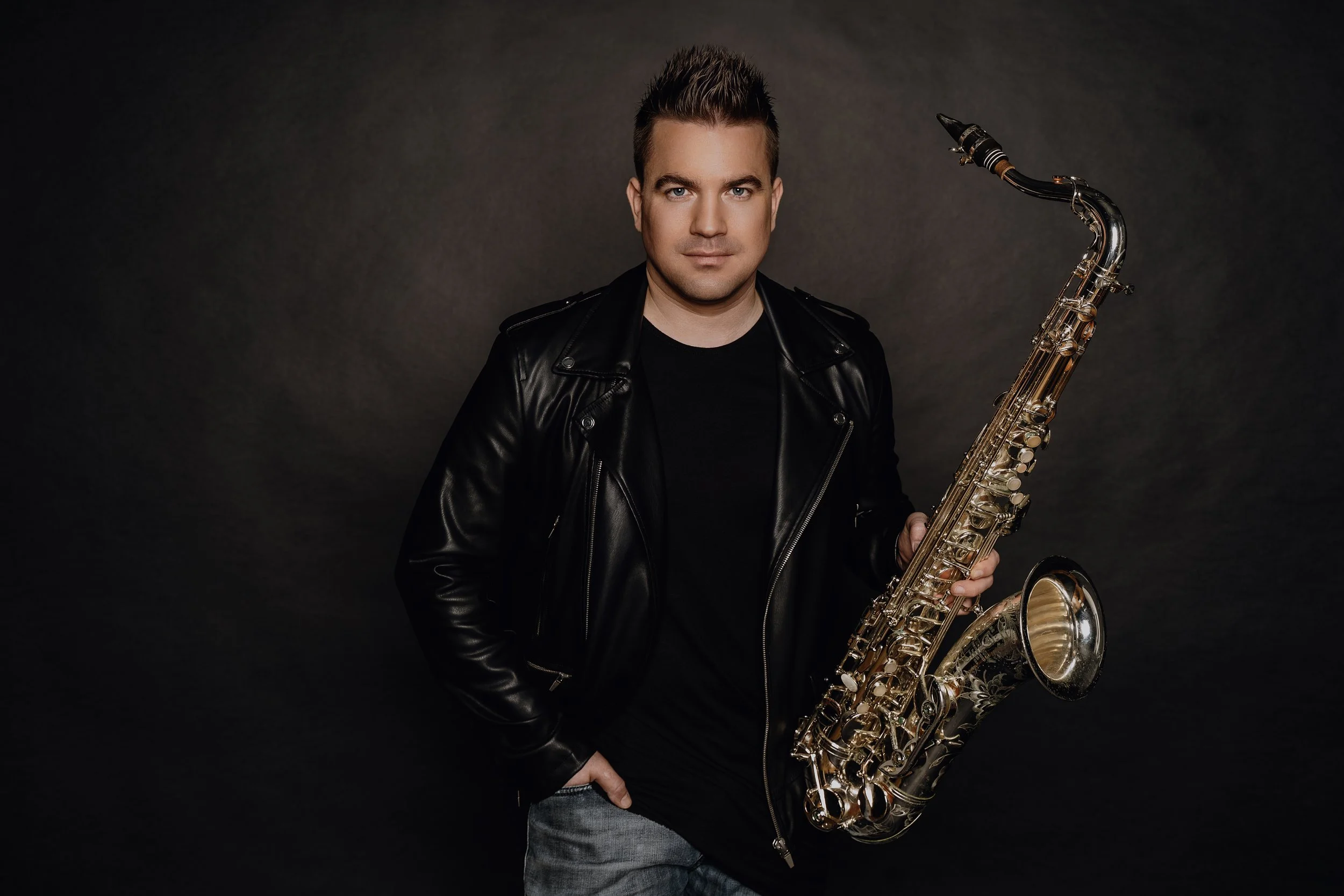 A young man with spiked hair wearing a black leather jacket holding a gold-colored saxophone against a dark background.