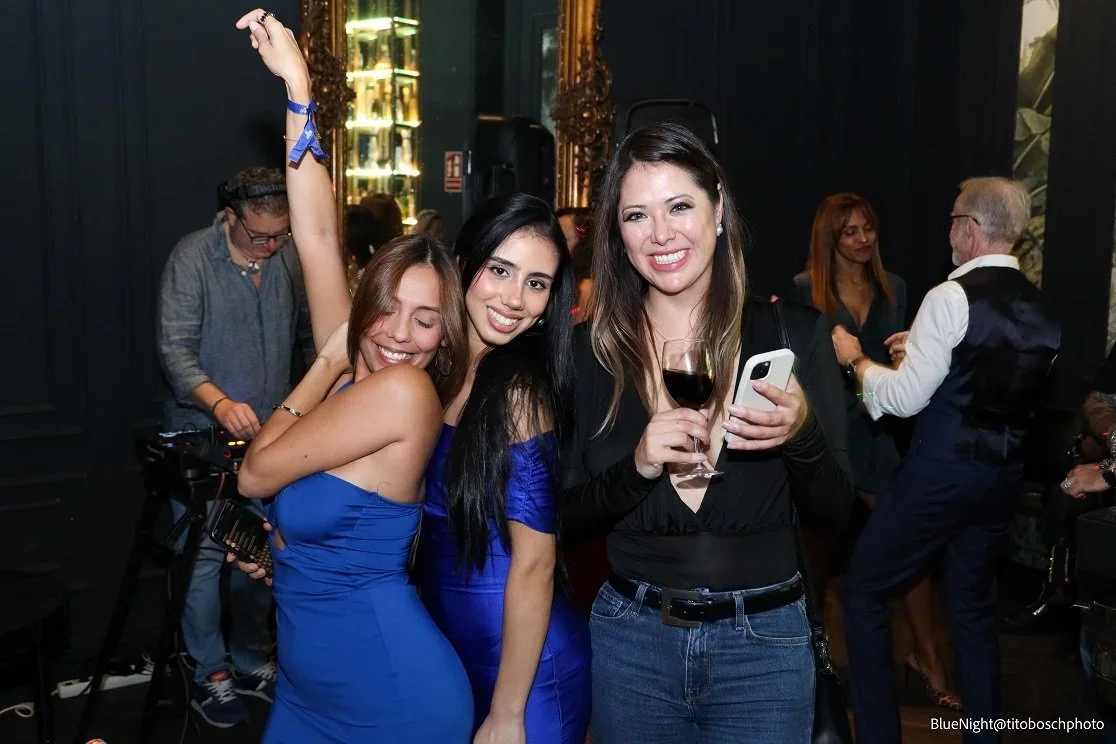 Three women at a social gathering, smiling and posing for a photo. They are holding drinks, and the woman on the right is using her phone. The background shows other people dancing and socializing.