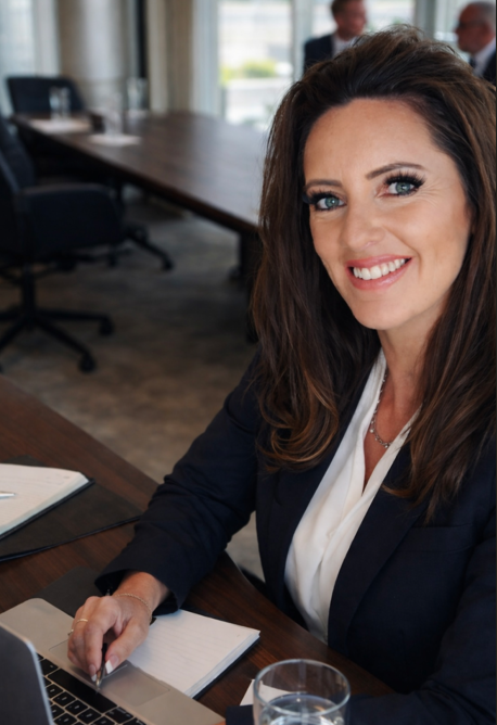 A woman with long brown hair smiling and sitting at a conference table in an office setting, with a laptop, notebook, and a glass of water in front of her. Two men are blurred in the background.