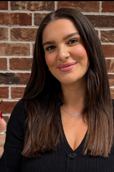 A woman with long brown hair and a smile, standing in front of a brick wall.