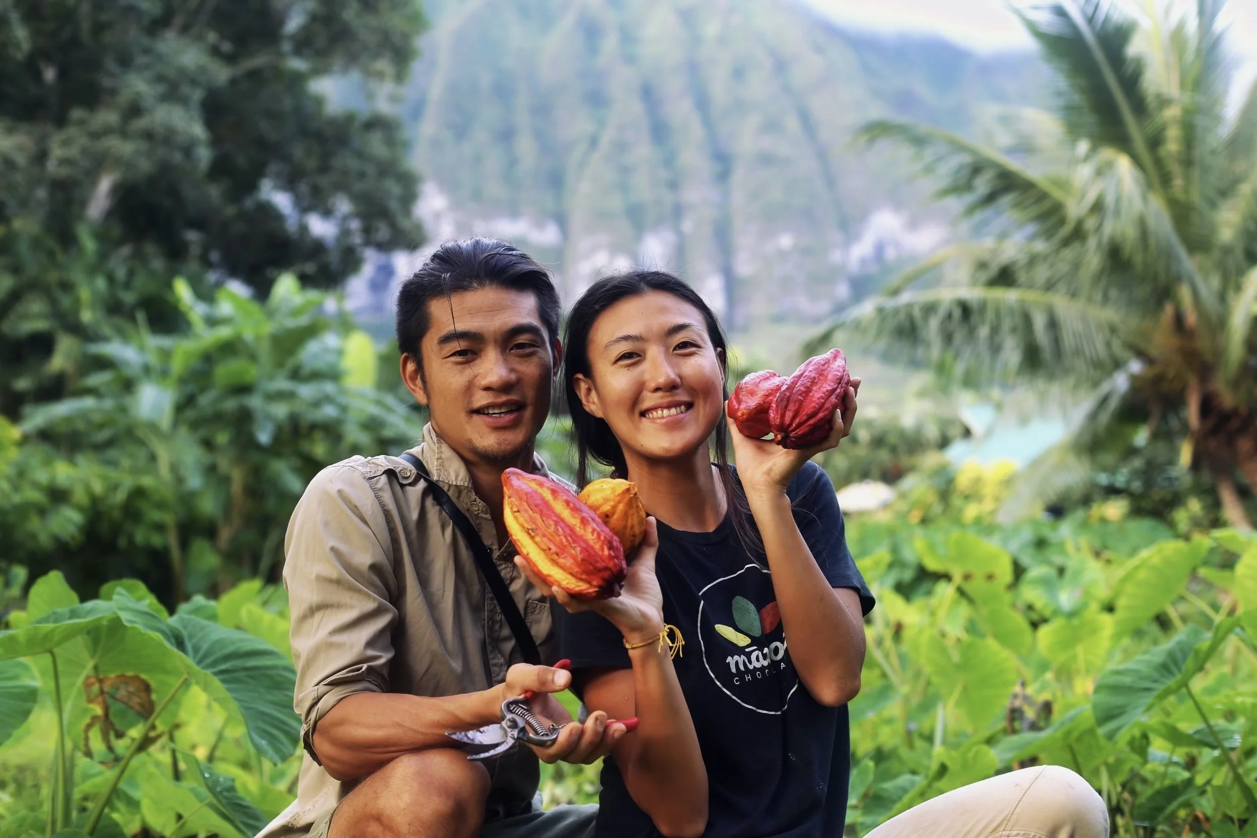 A smiling man and woman holding ripe cacao pods in a lush green jungle setting.