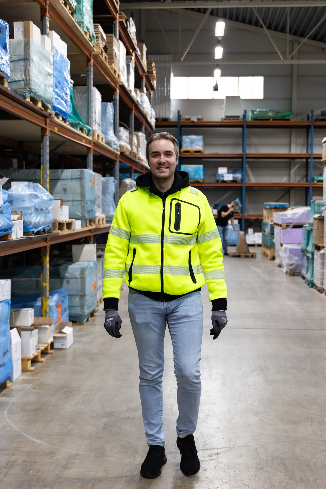 Nuori mies työasussa sisätiloissa varastossa, taustalla hyllyjä ja muita ihmisiä.
