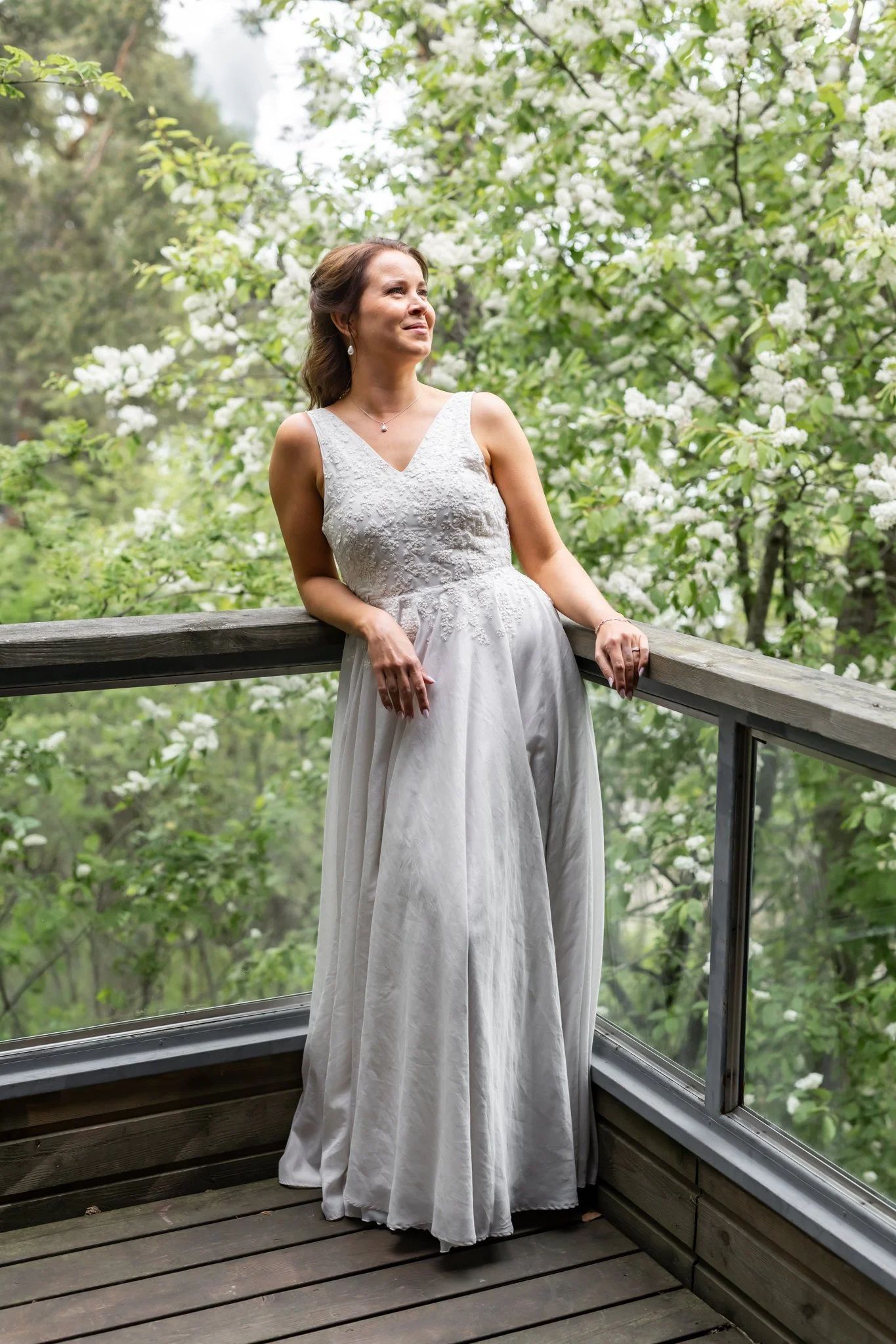 Nainen hääpuvussa seisoo terassilla, ympärillään vihreää ja valkoisia kukkia. Morsiamella on kaunis hääpuku.