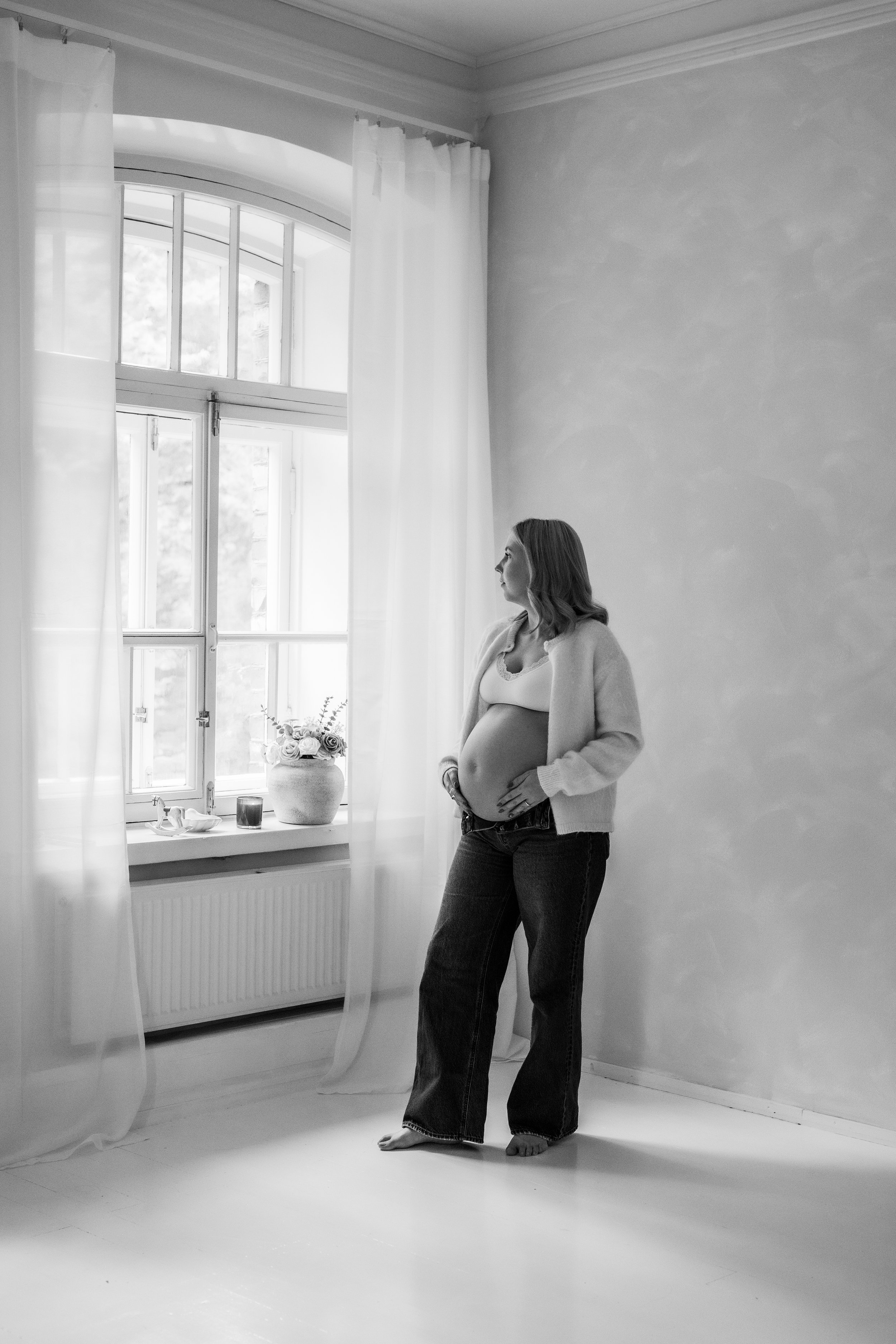 Raskaana oleva nainen seisoo ikkunan edessä hymyilemättä, katsellen ulos. Hän on pukeutunut vaaleaan puseroon ja farkkuihin, ja hänen kätensä ovat vatsallaan. Huoneessa on valkoinen seinä ja pehmeässä valossa.