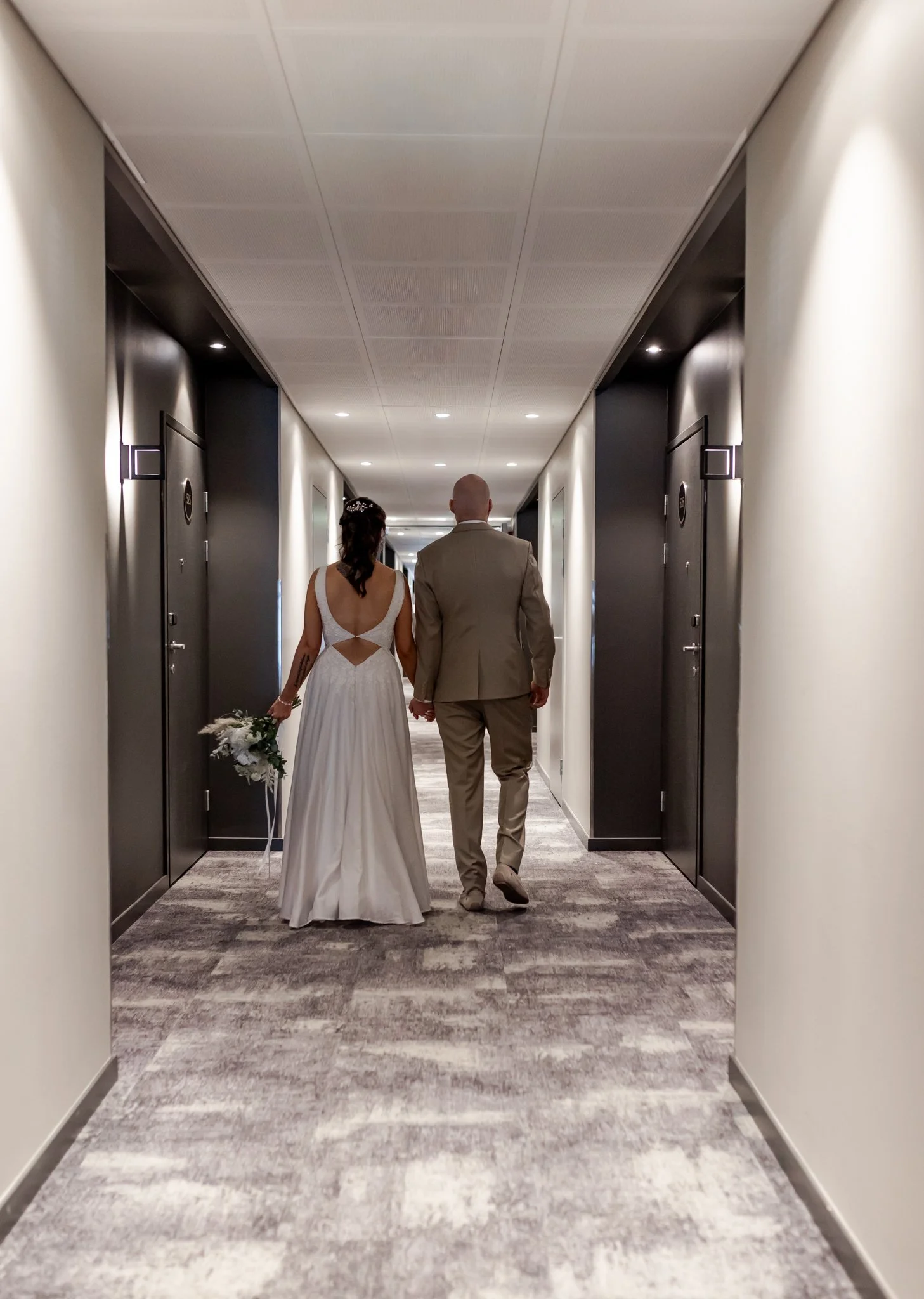 Hääpari kävelee käytävällä, naisella valkoinen hääpuku ja miehellä beige puku, nainen pitää kukkakimppua.