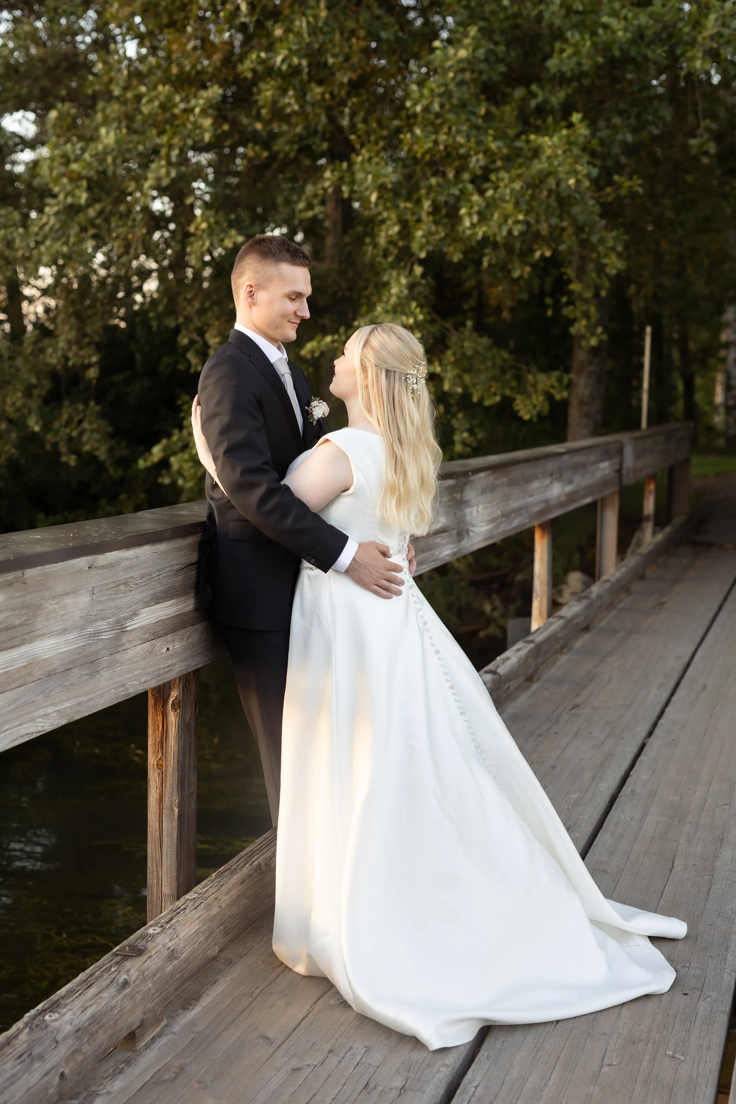 Hääpari seisoo toisissaan kiinni sillalla, taustalla puita ja vesi, iltavalaistus, nainen valkoisessa häämekossa ja mies mustassa pikkutakissa.