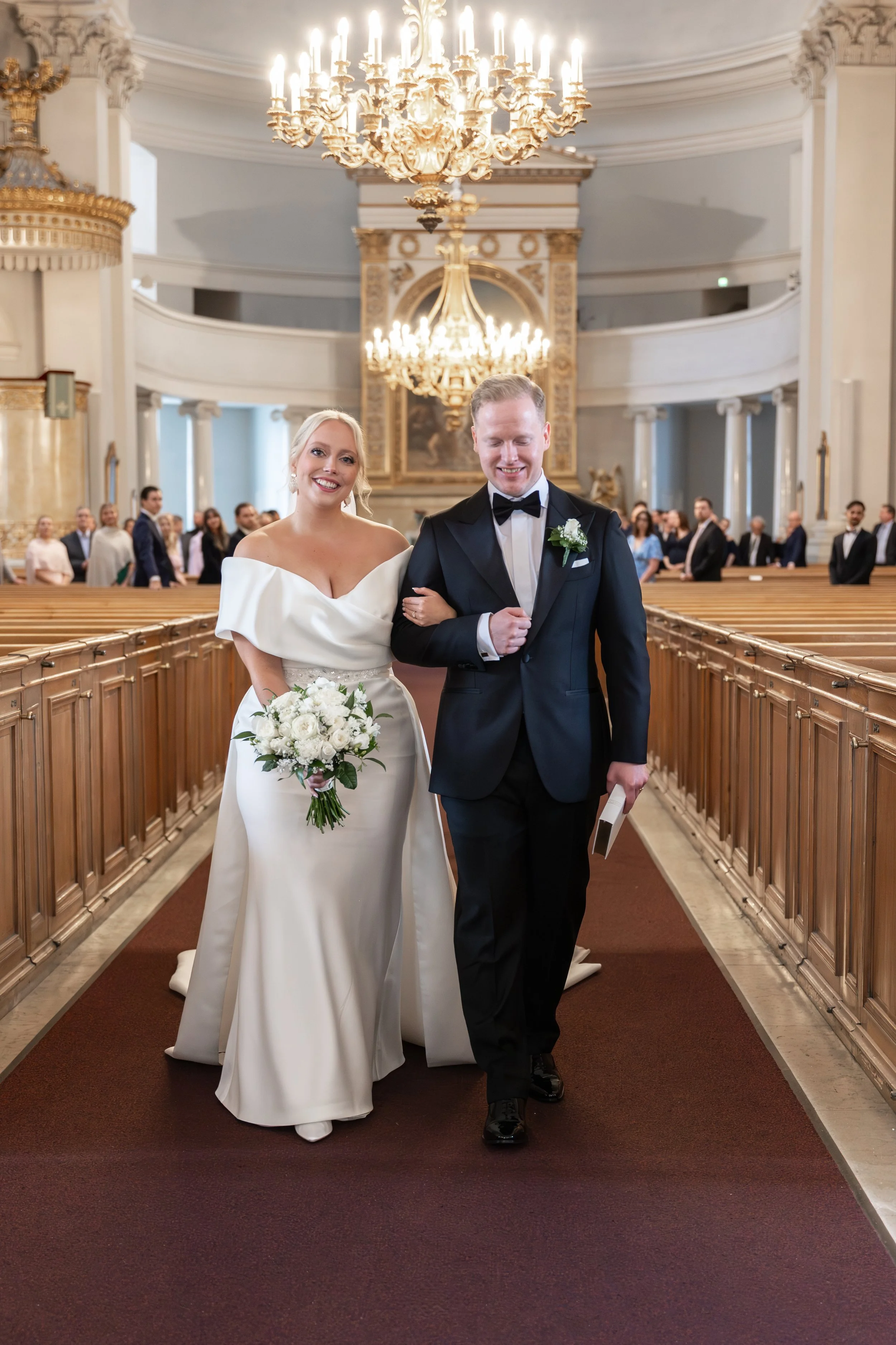 Hääpari kävelee kirkkosalin keskellä, sulhanen pitää morsianta kädestä ja heidät on juuri vihitty. Morsiamella on kaunis hääpuku ja huntu.