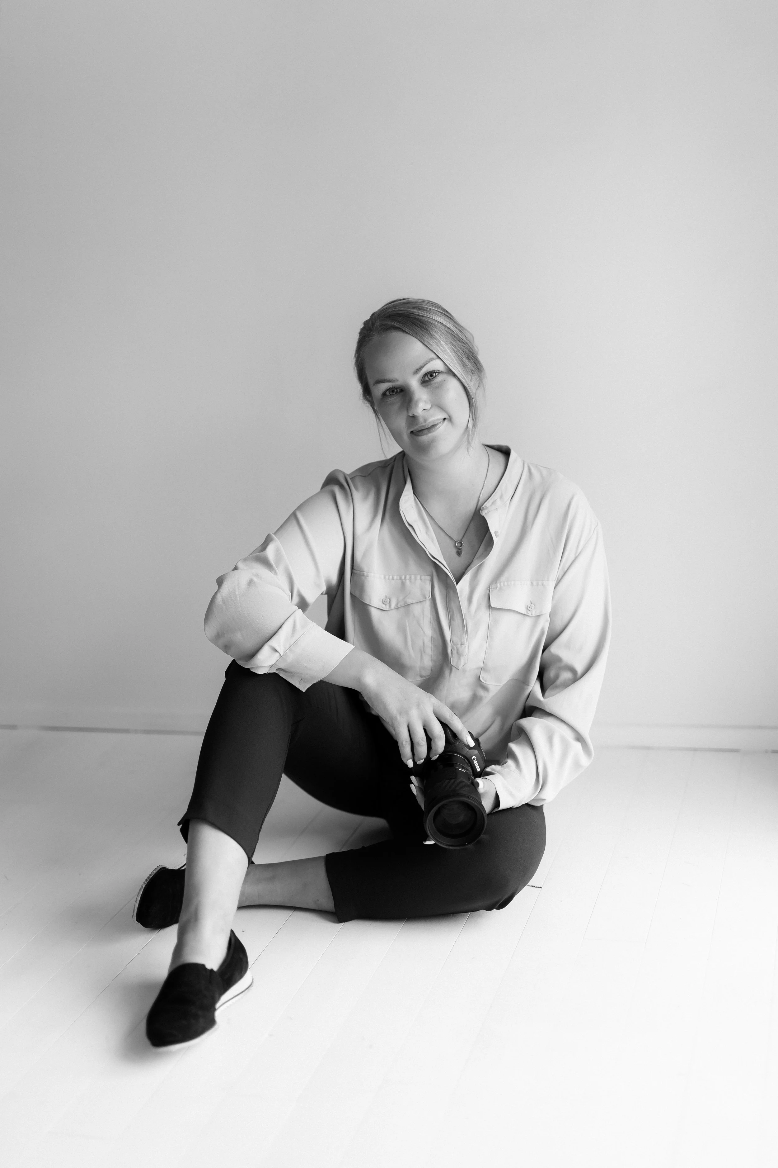 Mustavalkoinen kuva naisesta istumassa lattialla, hänellä on kamera kädessään, pukeutuneena valkoiseen paitaan ja mustiin housuihin.