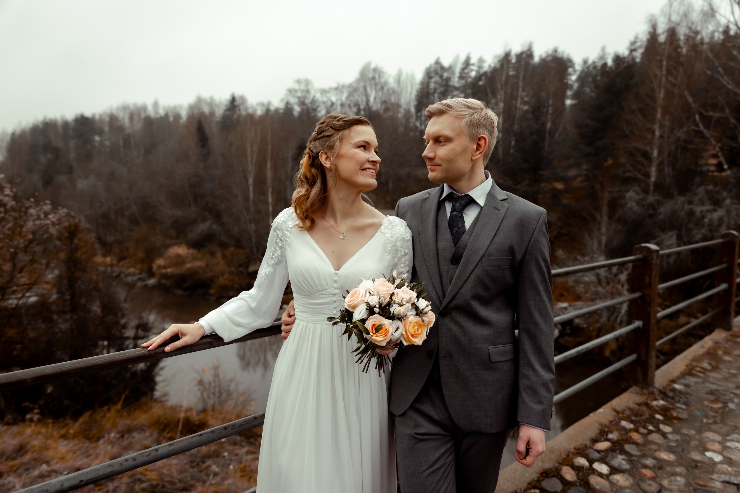 Hääpari seisoo sillalla ja katsoo toisiaan, morsian pitää kaunista kukkakimppua ja hääpari hymyilee toisilleen.