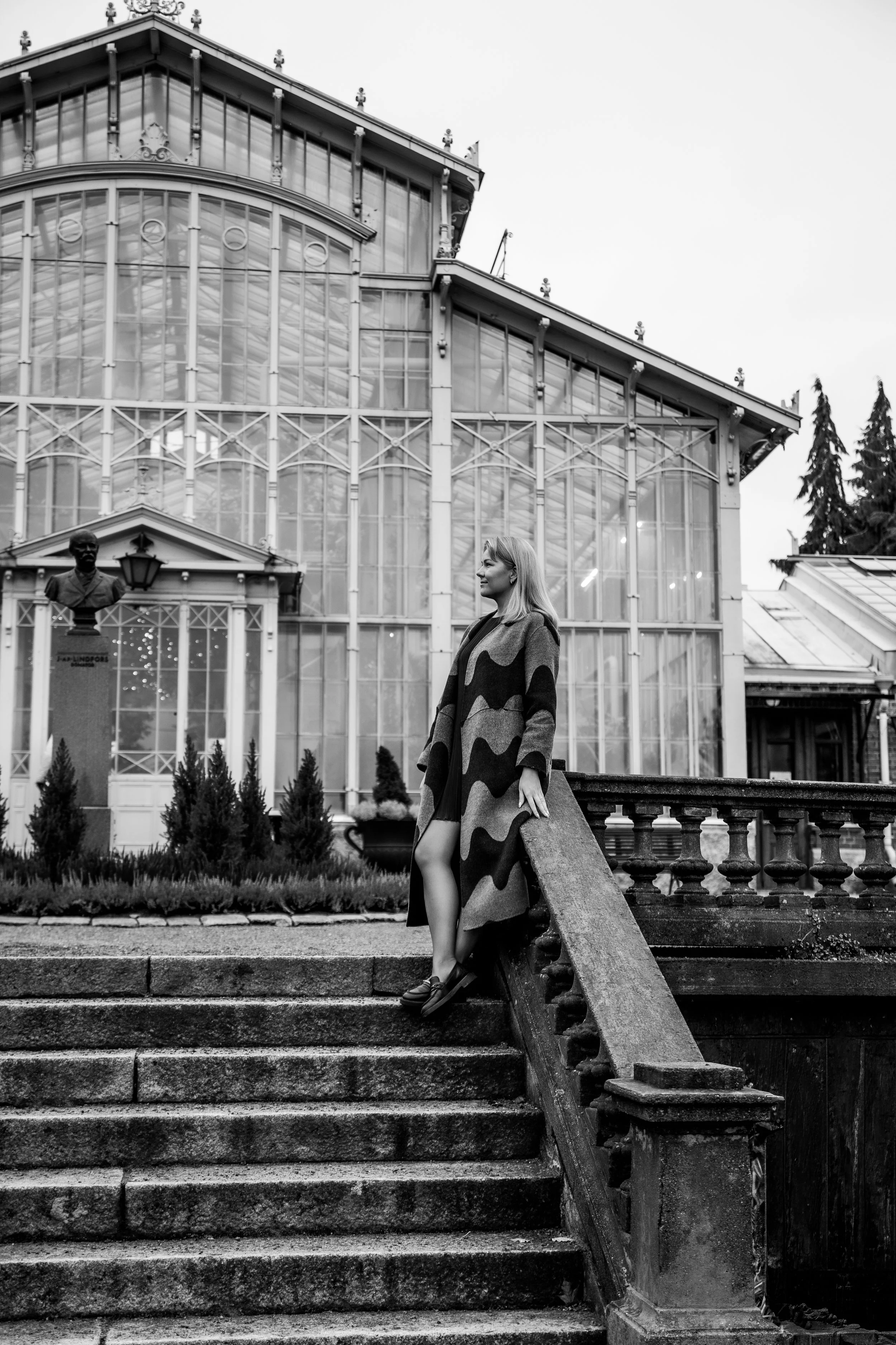 Nainen seisoo portaikossa ja taustalla iso ja upea lasinen talo. Mustavalkoinen kuva.