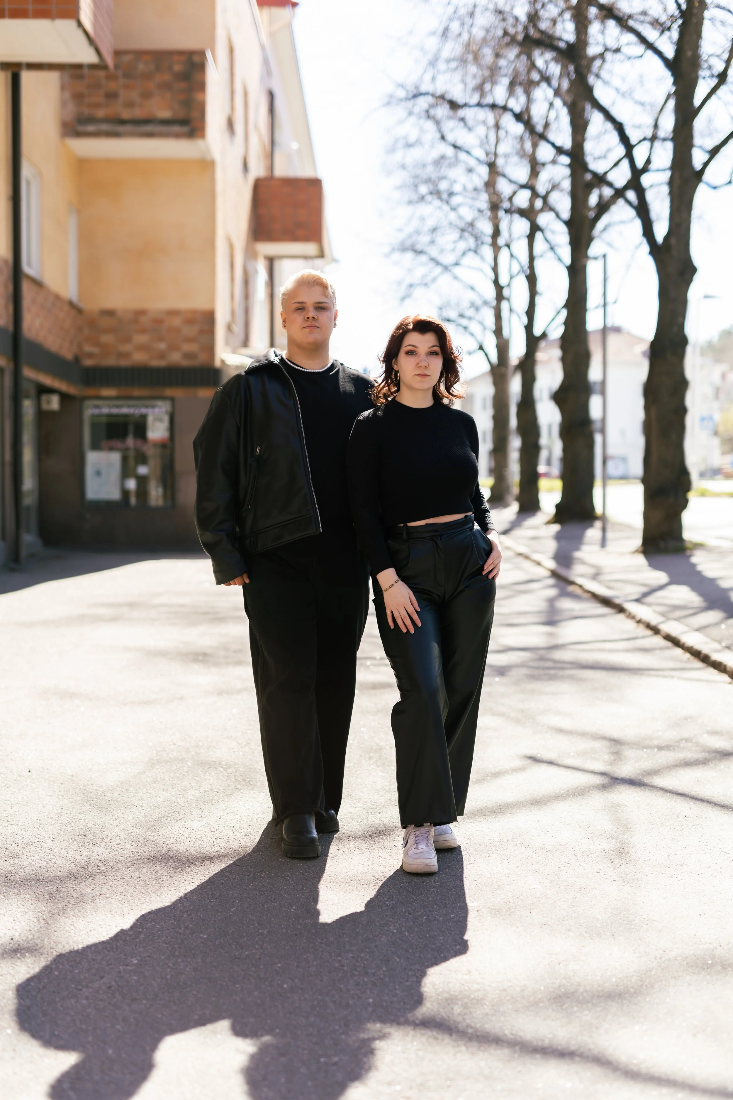 Kaksi nuorta aikuista kävelee kaupunkimaisemassa, taustalla rakennuksia ja puita, aurinko paistaa voimakkaasti.