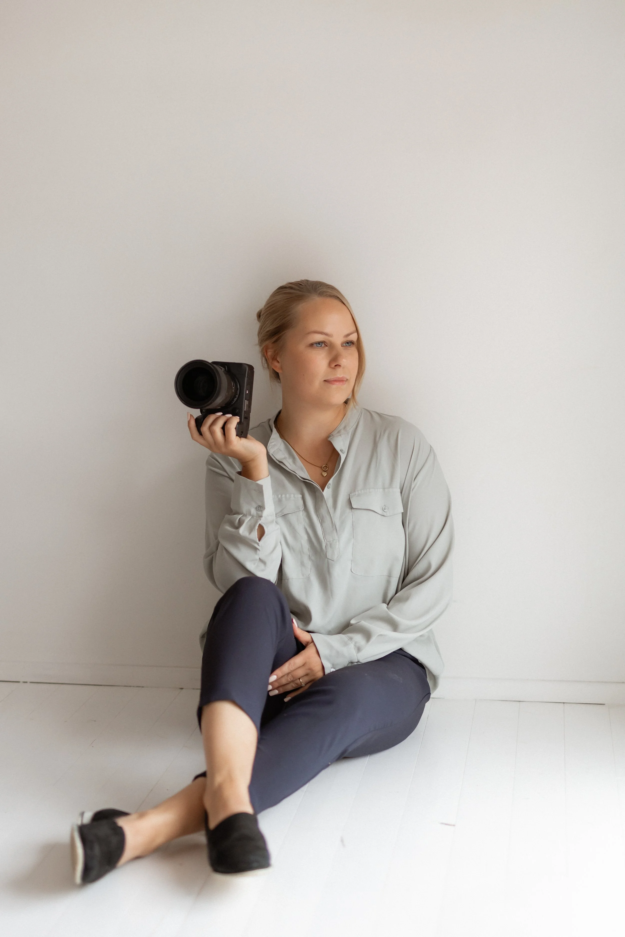 Nainen istuu maassa, pitää kameraa kädessään ja katsoo sivuun, taustalla valkoinen seinä.