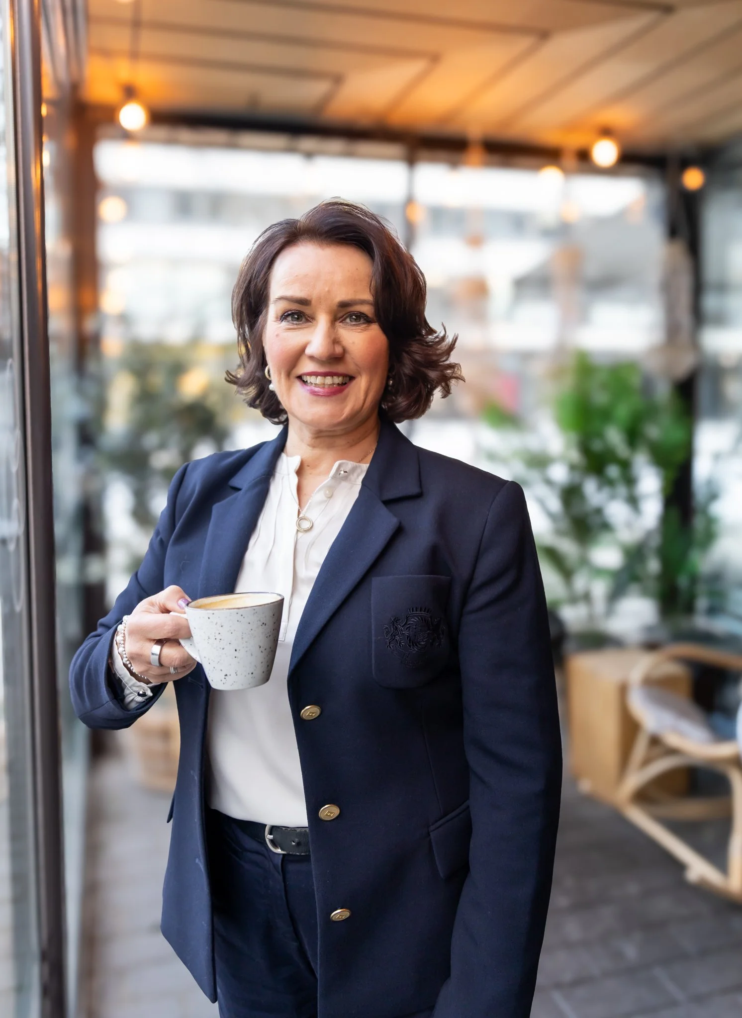 Kaunis nainen hymyilee kahvikuppi kädessä. Naisella on päällään tumma jakkupuku. Henkilökuva.