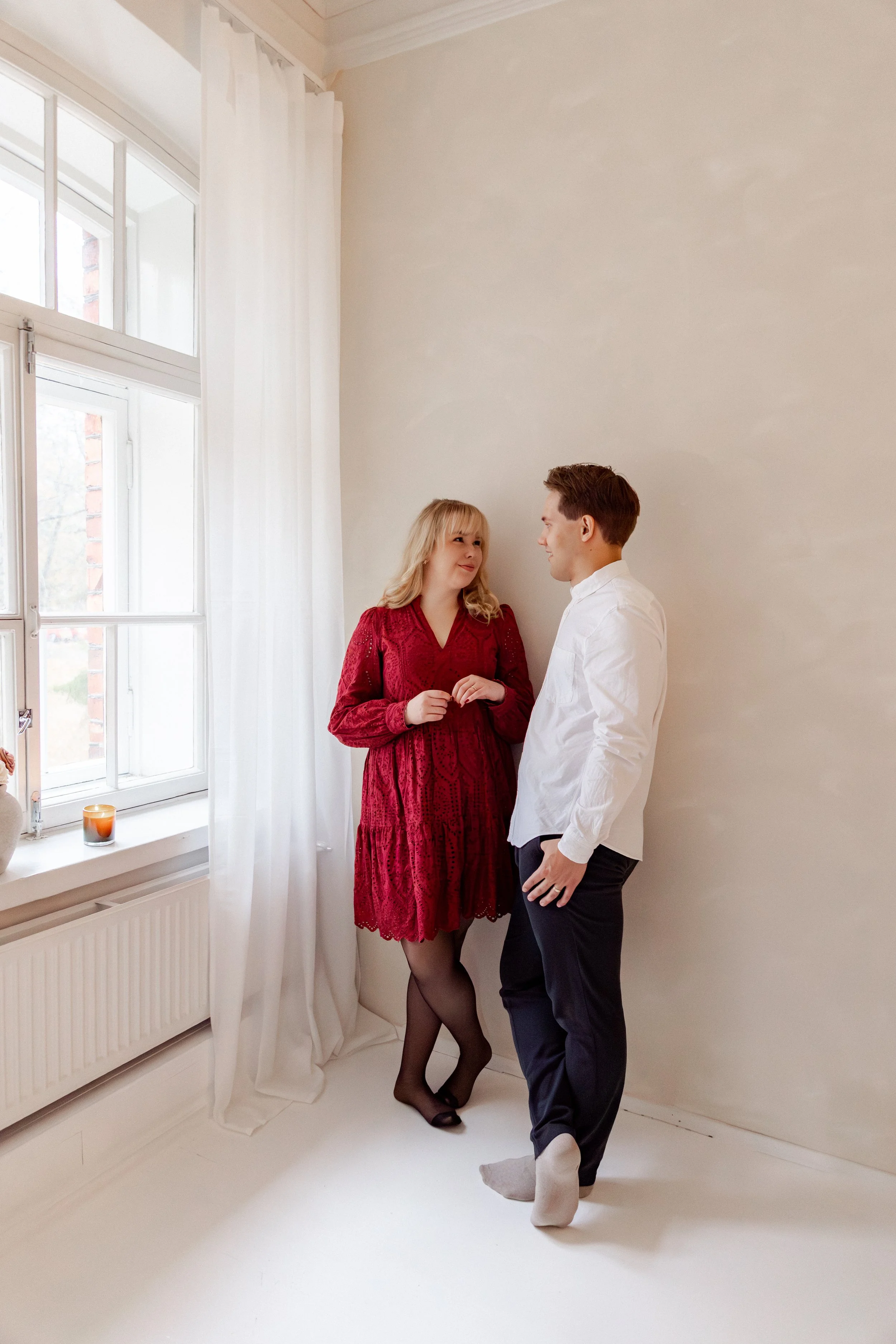 Kaksi ihmistä seisoo yhdessä huoneessa, nainen pukeutuneena punaisiin mekkoihin ja mies valkoiseen pikkutakkiin, he katsovat toisiaan ja hymyilevät.