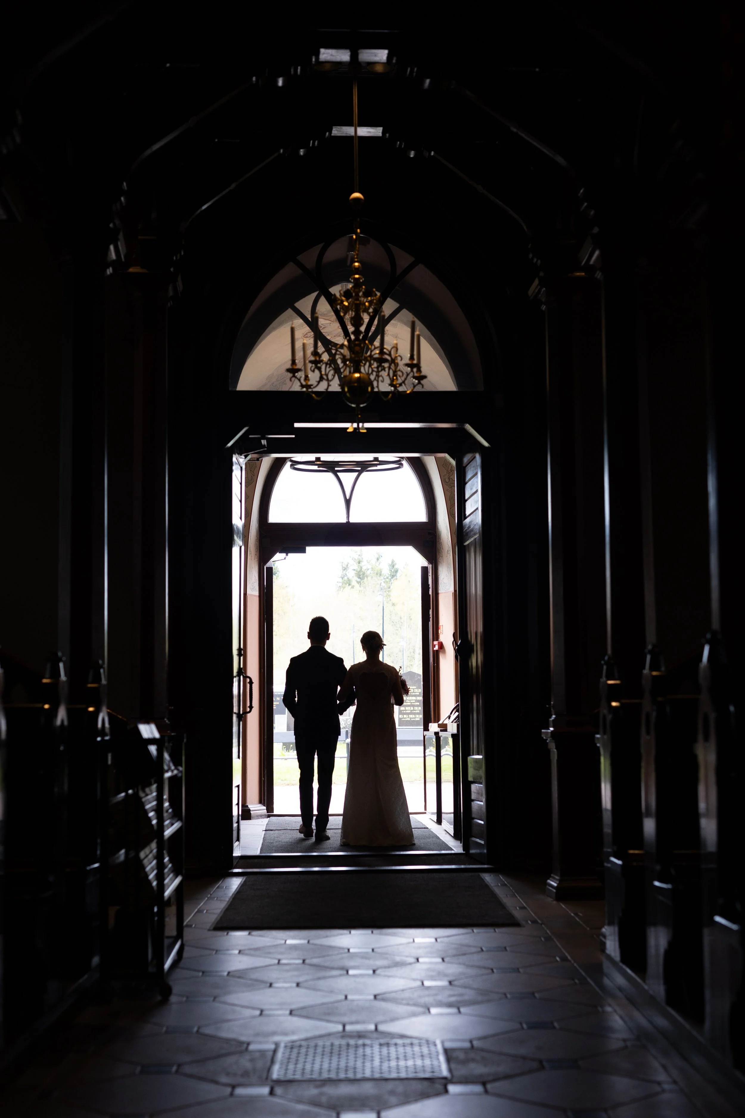 Hääpari kävelemässä käsi kädessä vihkikirkosta ulos. Kuva on tumma ja hääparista näkyy kaunis siluetti.