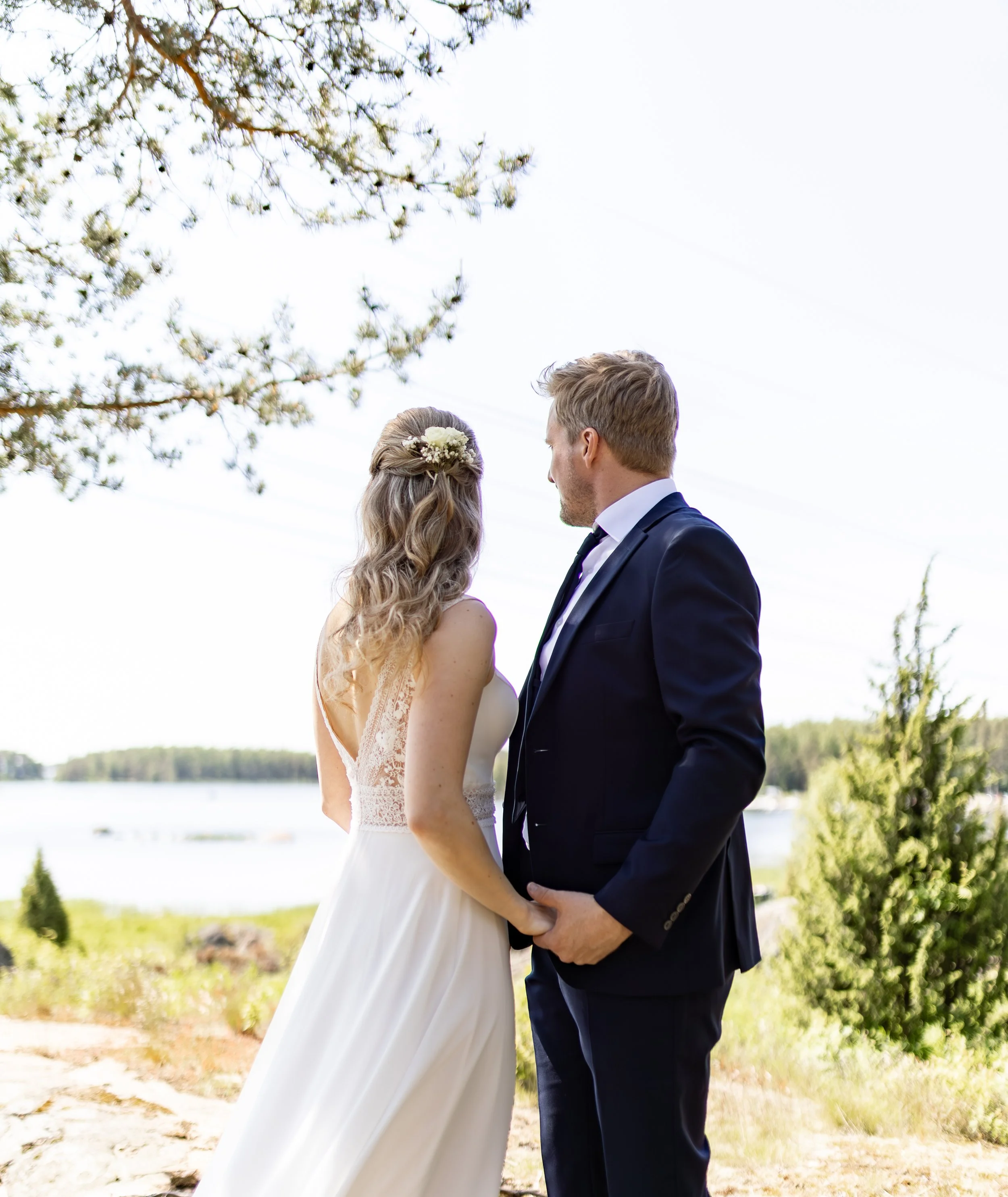 Hääpari seisoo lähikuvassa rakastavasti toisistaan käsi kädessä ulkona, taustalla järvi ja puut. Morsian pitää sulhasesta hempeästi kiinni.
