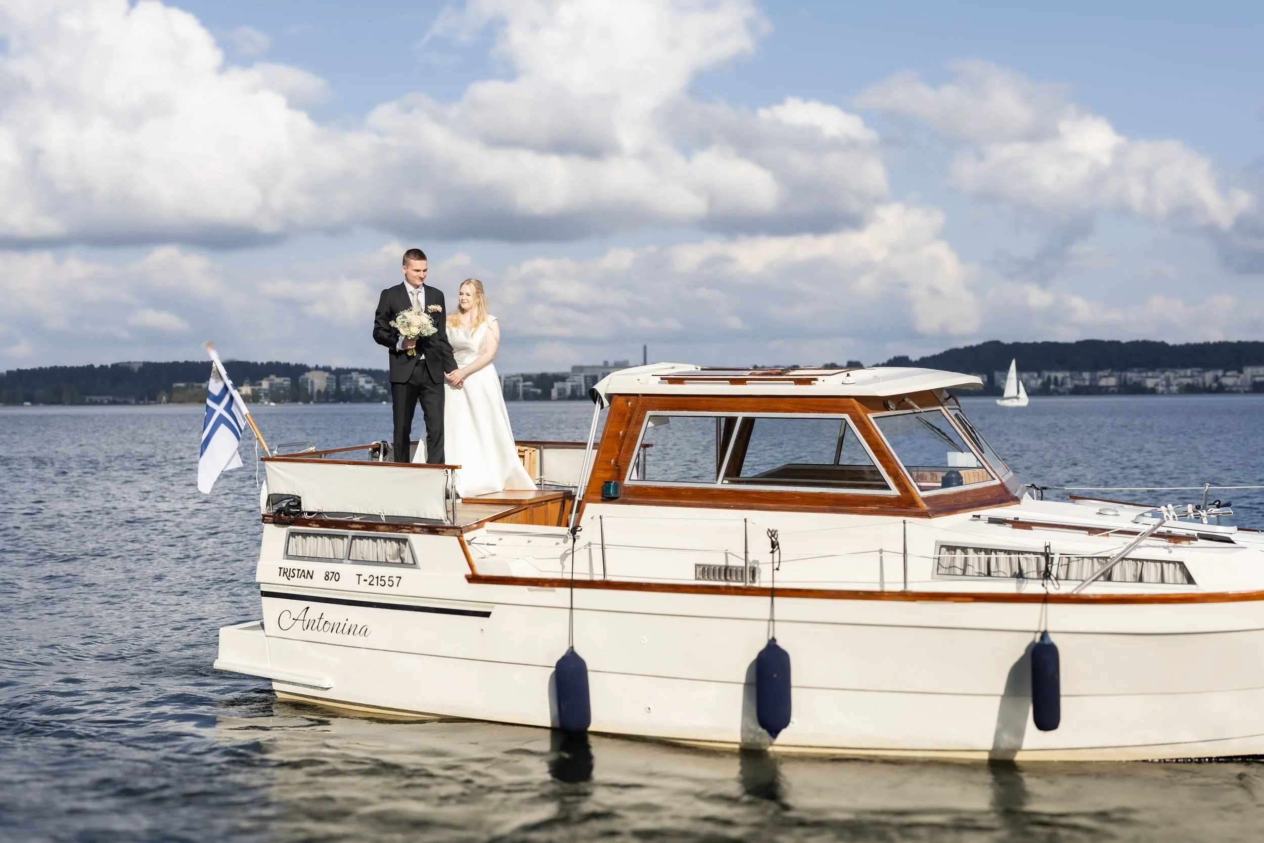 Hääpari seisoo veneen keulassa, taustalla vesi ja pilvinen taivas.