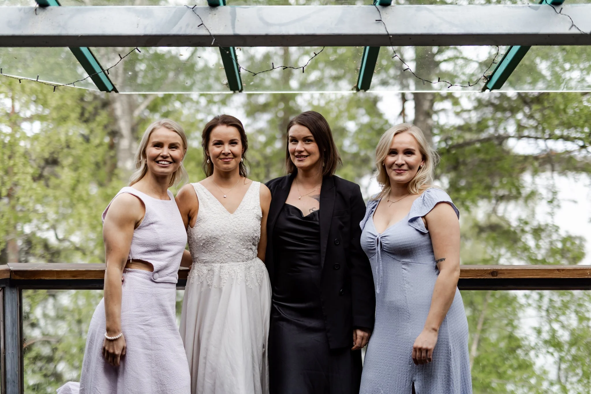 Neljän naisen ryhmäkuva ulkona, vihreitä puita taustalla, yksi naisista pukeutunut hääpukuun.