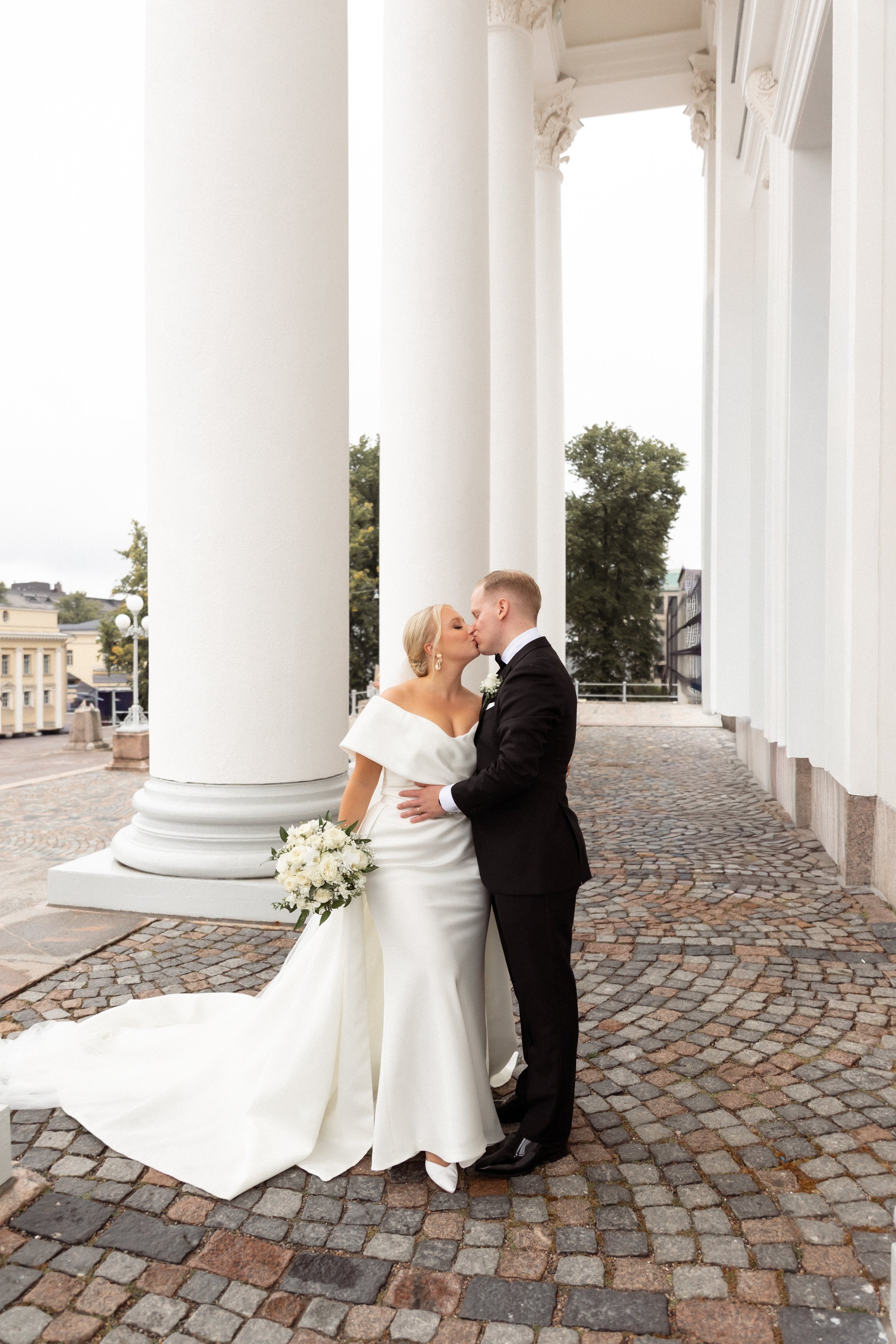 Hääpari suutelee toisiaan Helsingin tuomiokirkon edessä. Morsiamella on upea valkoinen hääpuku ja iso kukkakimppu. Sulhanen pitelee morsiantaan lähellä itseään.