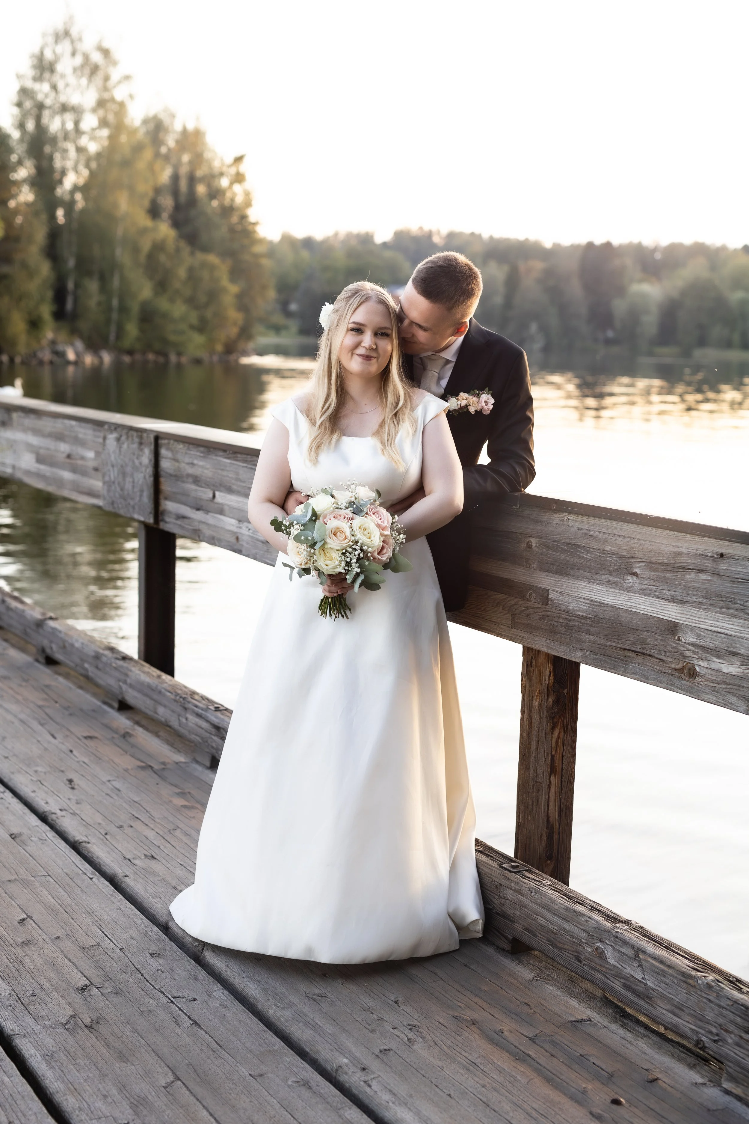 Hääpari järven rannalla puusillalla, nainen valkoisessa hääpuvussa ja mies mustassa puvussa, nainen pitää kukkakimppua, taustalla vesi ja puita.