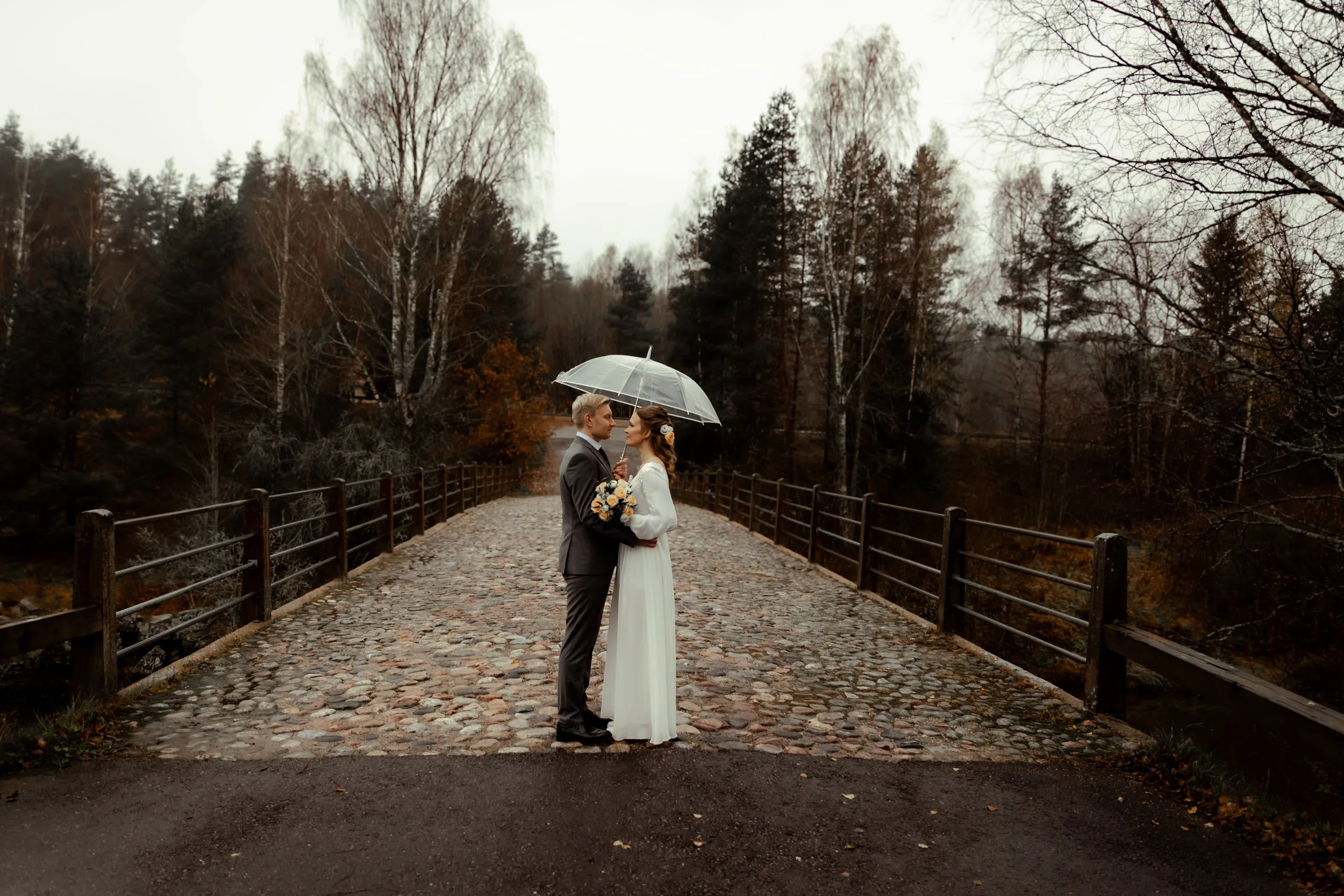 Hääpari seisoo sateenvarjon alla keskellä siltaa. Hääpari suutelee toisiaan ja sää on sumuinen. Morsiamella on kaunis hääpuku. 