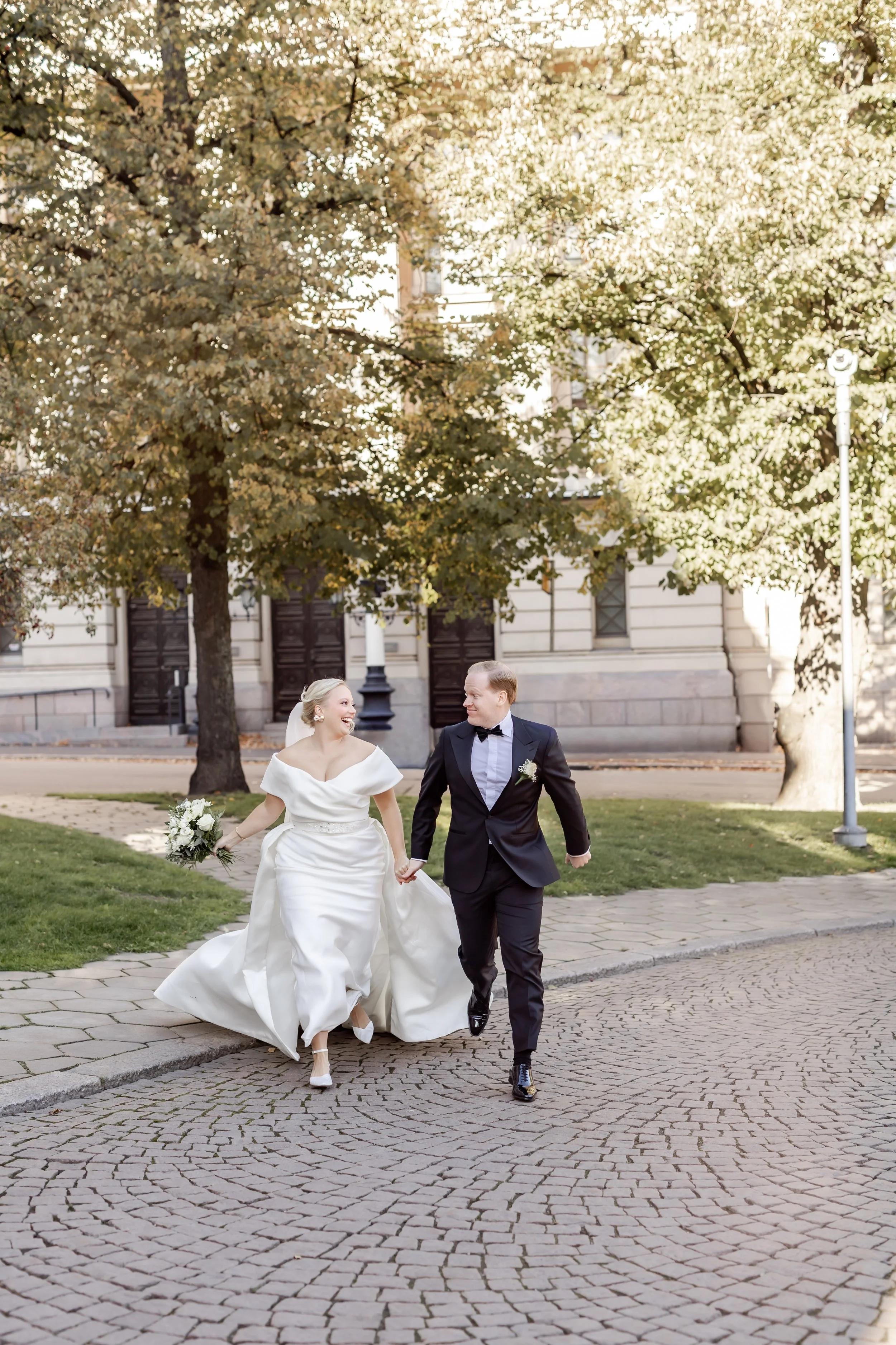 Hääpari kävelee käsi kädessä puistossa, nainen pukeutuneena valkoiseen häämekkoon ja mies pukuun, takana puiden ja vanhan rakennuksen näkyvät.