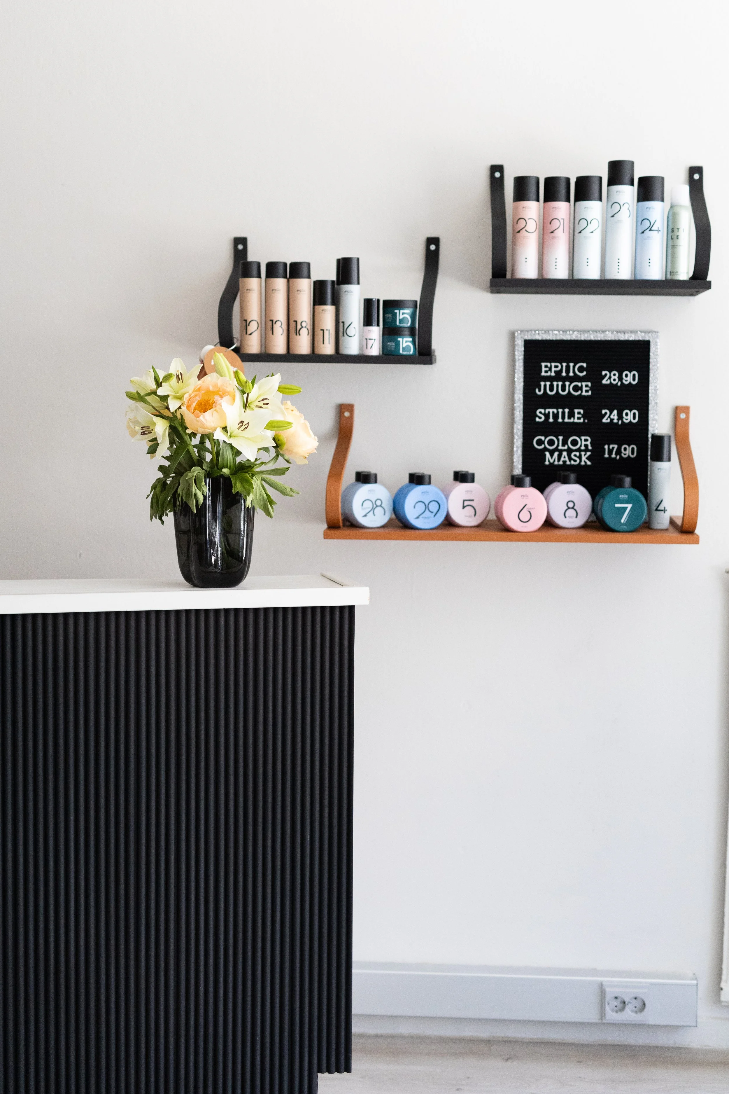 Kukka-asetelma mustassa maljakossa pöydällä, taustalla seinällä hyllyt ja valkoinen seinä.