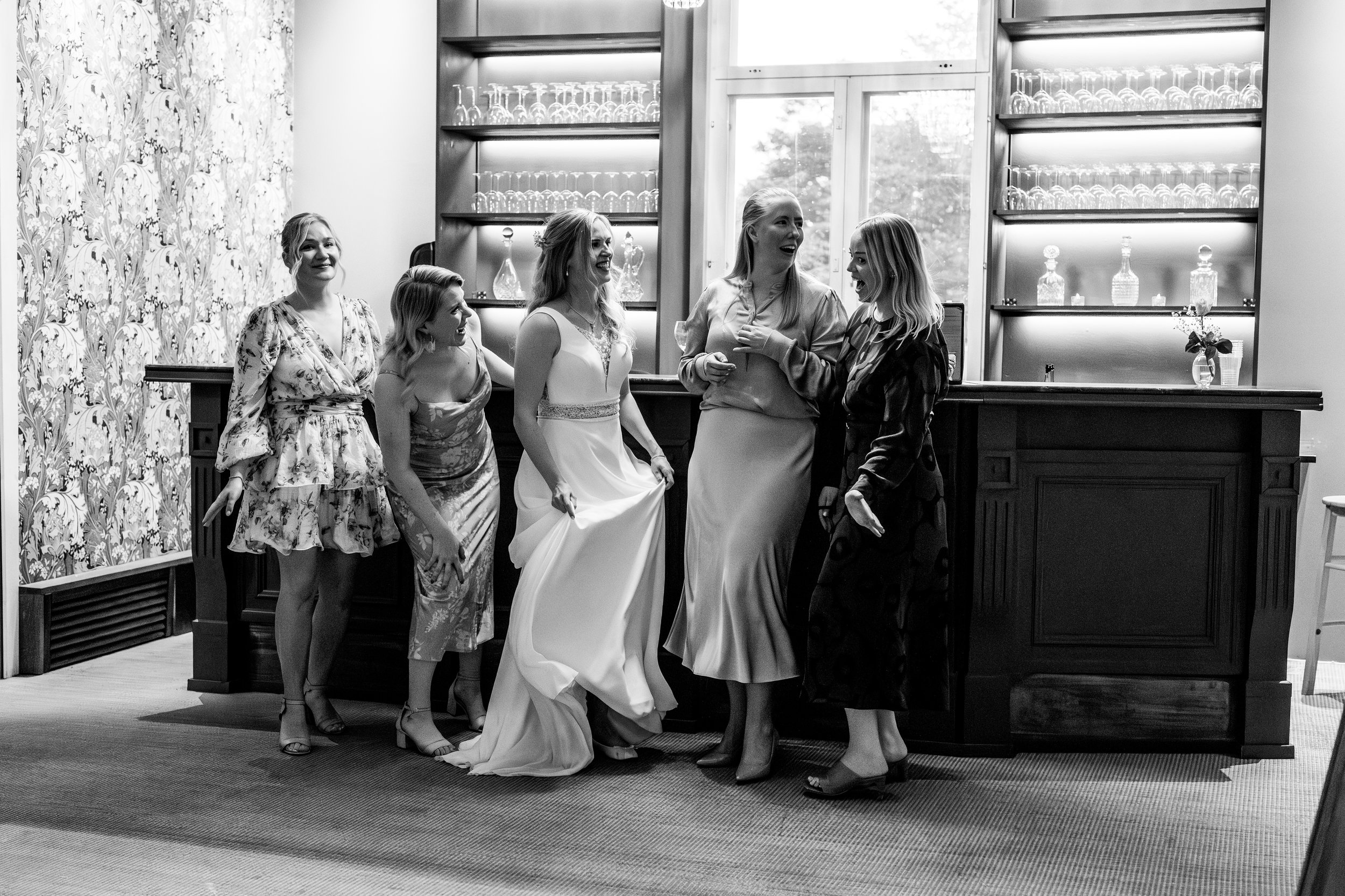 Kuvassa on kuusi naista, jotka ovat pukeutuneina juhlamekkoihin ja ovat iloisia ja nauravaisia, seisomassa baarin äärellä, taustalla hyllyt, joissa on laseja ja koriste-esineitä. Keskellä morsian joka heiluttaa kaunista mekkoaan. 