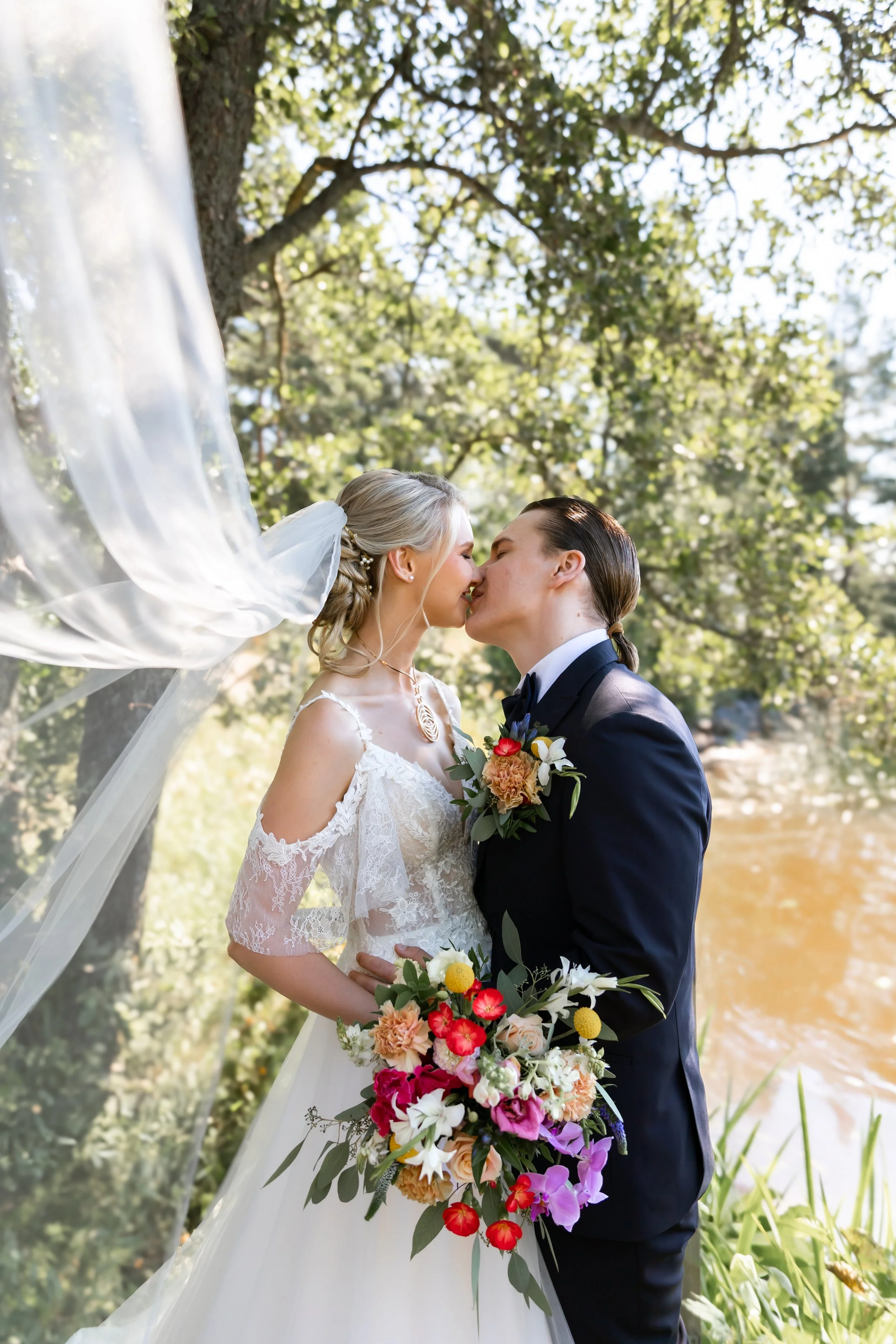 Hääpari suudellessaan luonnon helmassa. Morsiamella kaunis hääpuku ja huntu joka hulmuaa tuulessa. Kaunis värikäs kukkakimppu.