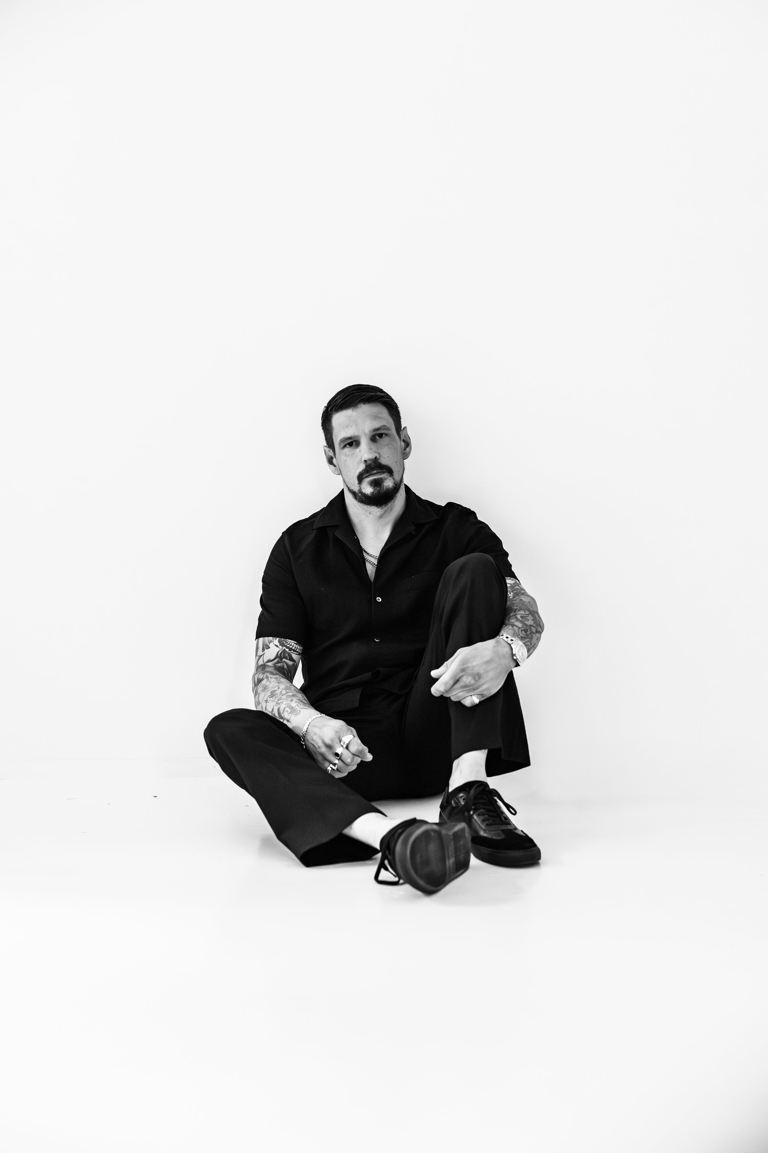 Henkilö istuu lattialla valkoisella taustalla, pukeutuneena mustaan pukuun, ja hänellä on tatuointeja ja rannekello.