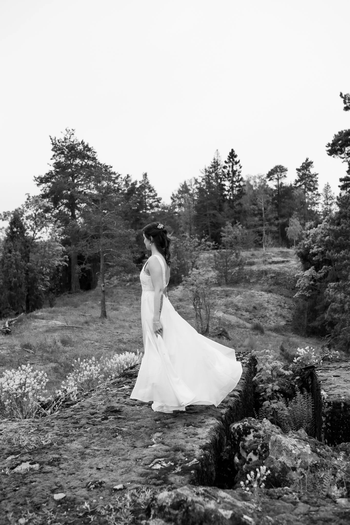 Morsian seisoo upeassa hääpuvussa kalliolla. Morsian pyörähtää ympäri ja helma hulmuaa kauniisti. Kuva on mustavalkoinen ja filmikuvatyylinen.