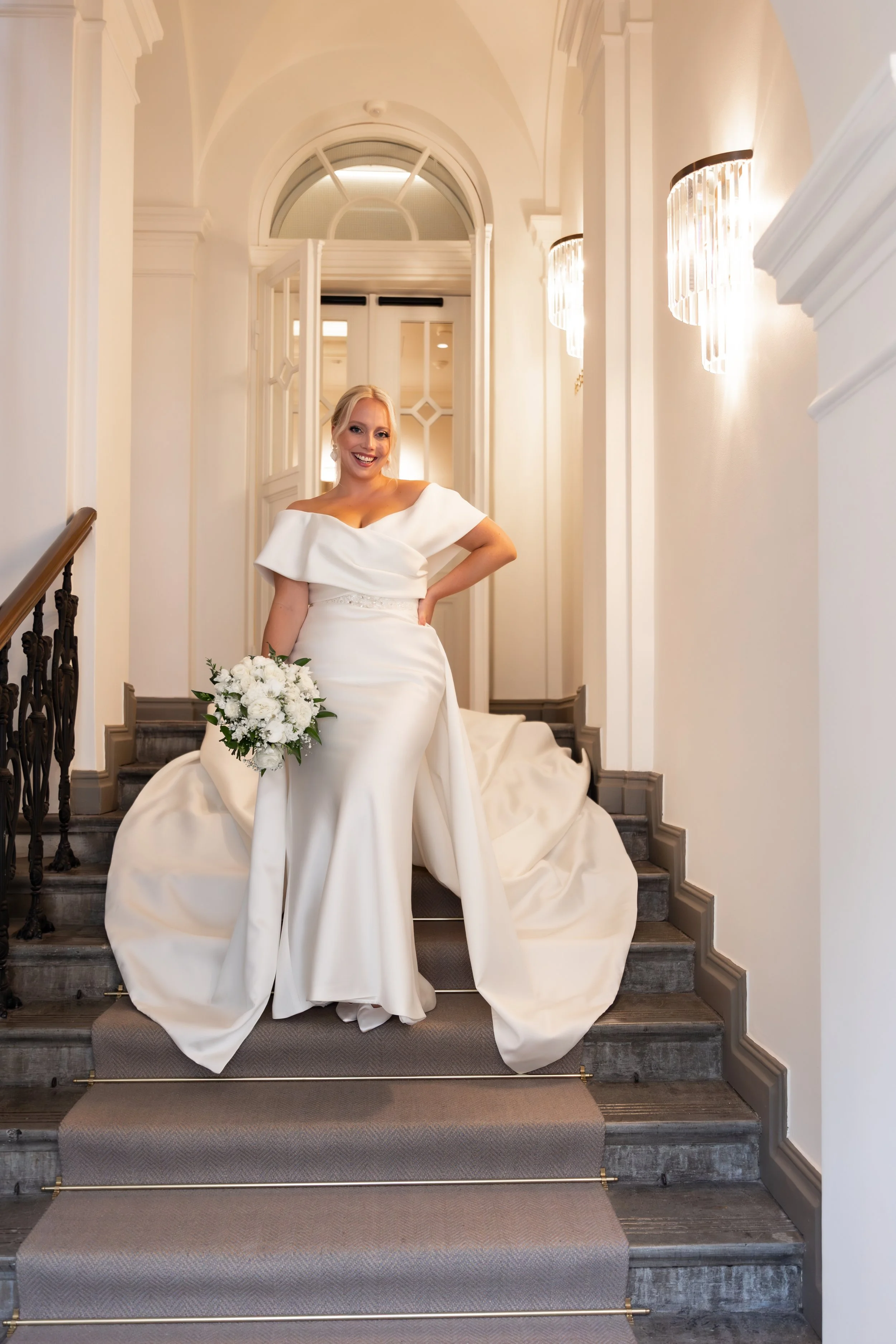 Kaunis morsian seisoo hotellin portaikossa. Hänellä on upea hääpuku ja kukkakimppu. Morsian hymyilee leveästi.