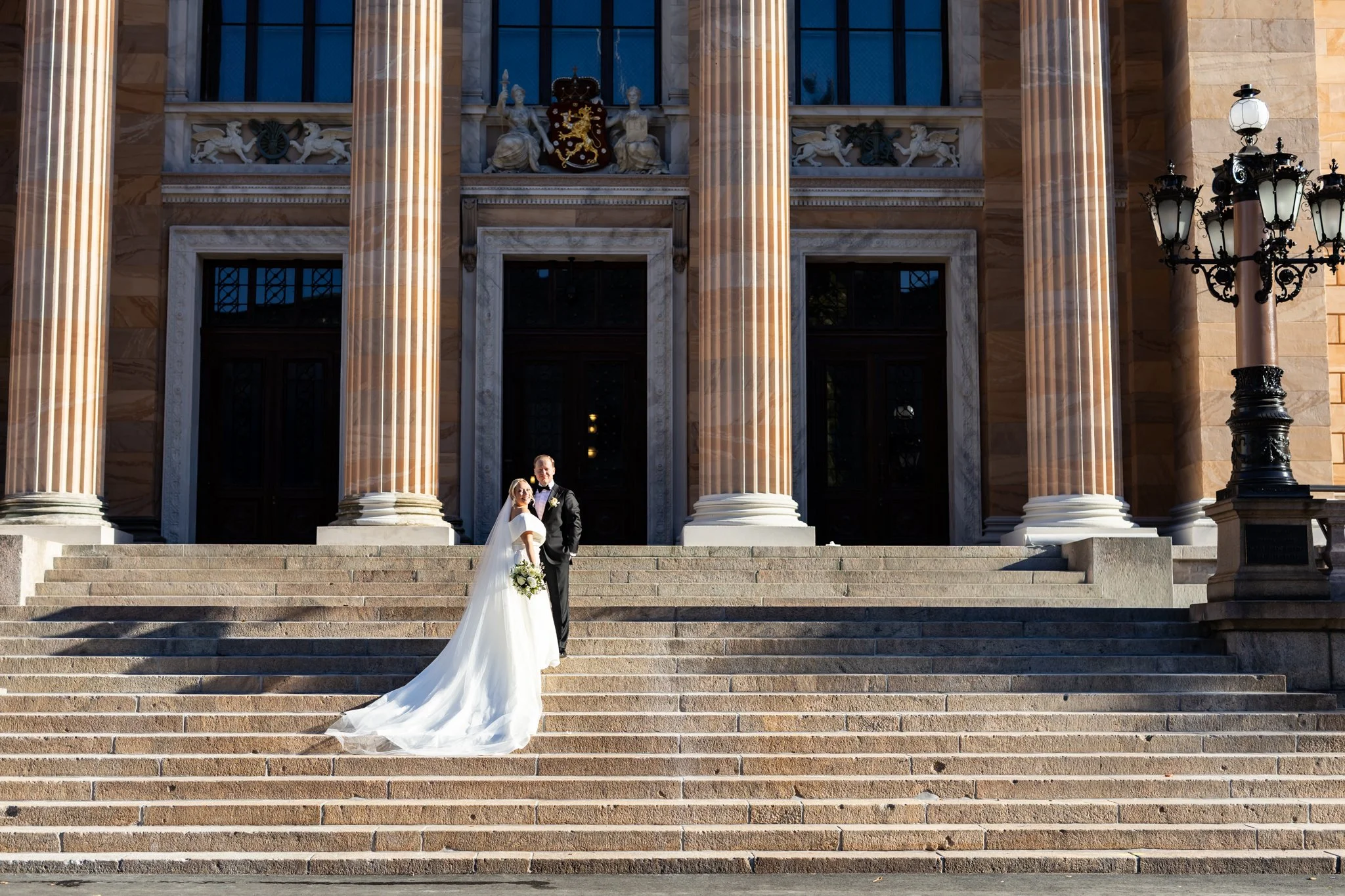 Hääpari seisoo kauniin rakennuksen edessä Helsingissä. Morsiamella kaunis hääpuku jossa pitkä laahus.
