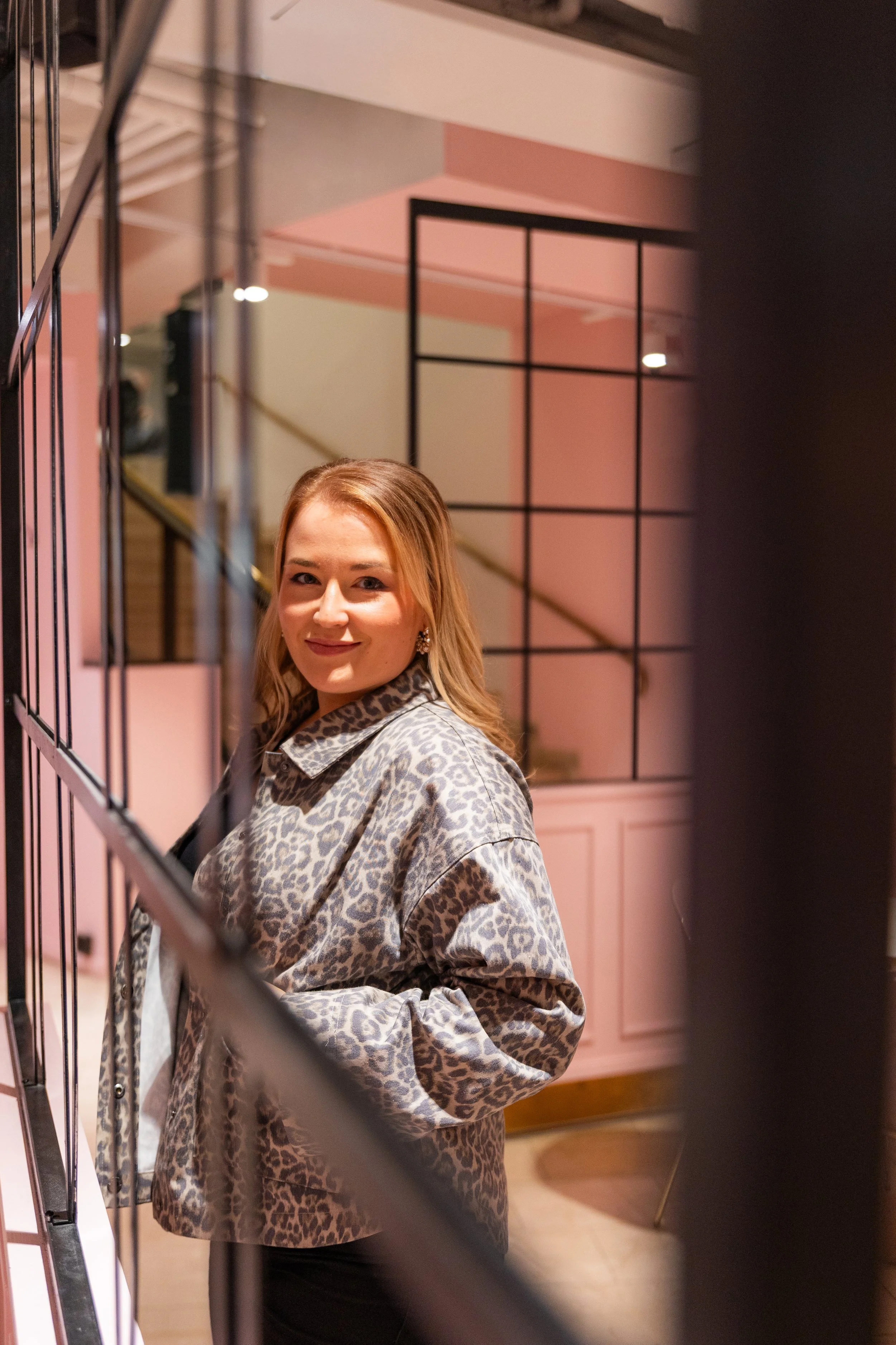 Nainen katsoo kameraan sisätiloissa, taustalla vaaleanpunainen ja valkoinen sisustus ja seinä, värit ja muotoilut modernit.