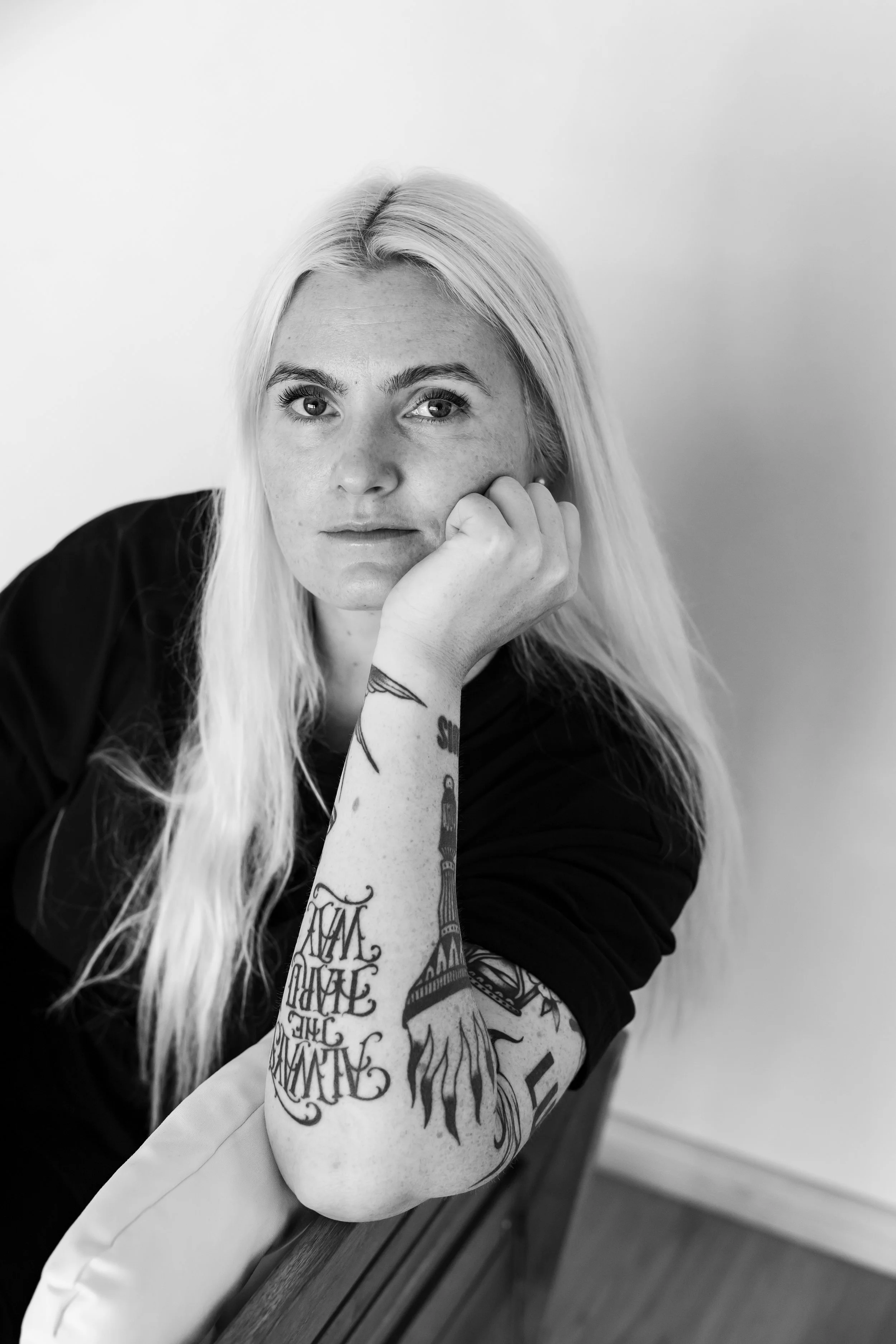  Nainen istuu tuolilla ja katsoo kameraan. Naisella on vaaleat hiukset ja käsissä paljon tatuointeja. 