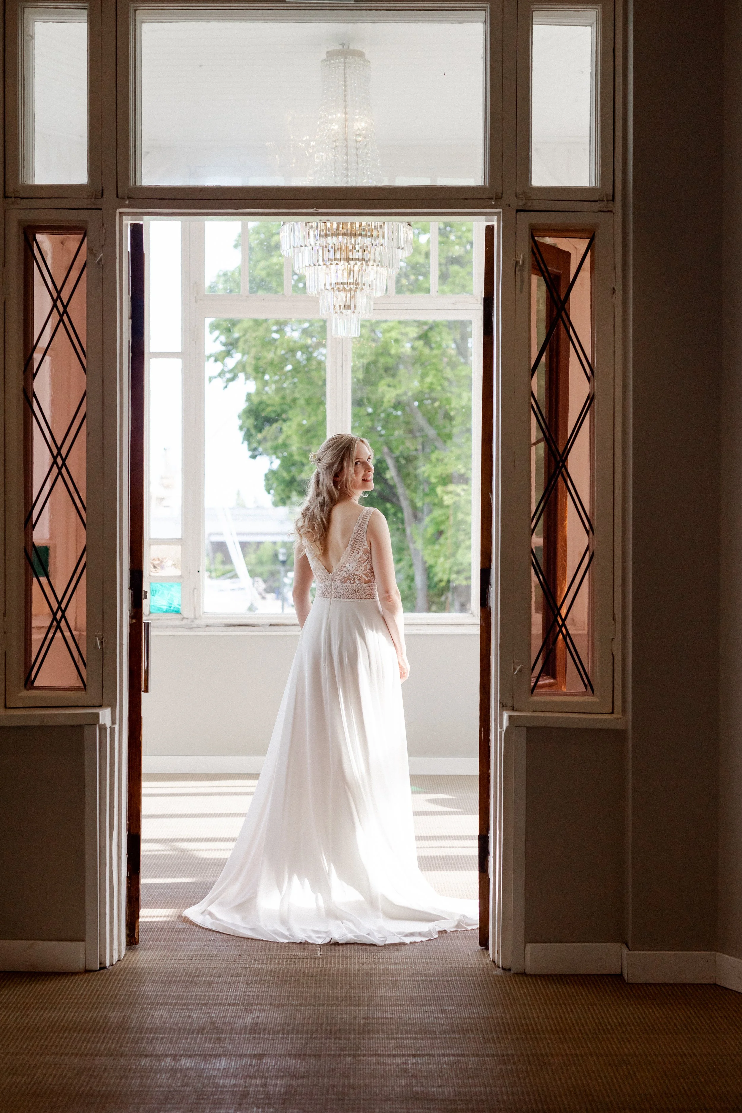 Nainen häämekossa seisoo sisällä katseen kääntyessä ikkunalle, taustalla vihreitä puita ja kristallikruunu katossa.