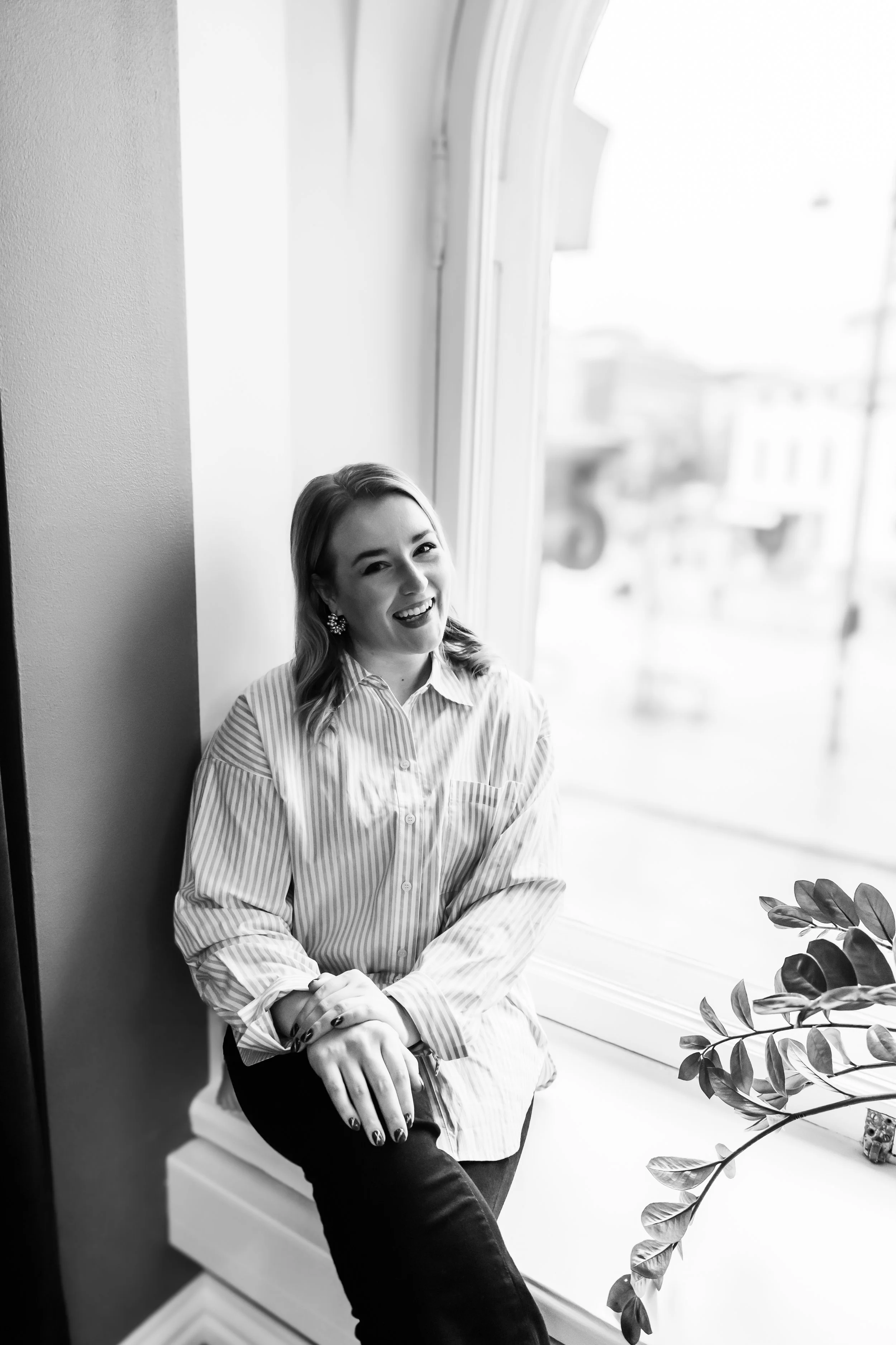 Nuori nainen istuu ikkunalaudalla sisällä hymyillen, hänellä on päällään raidallinen paita ja korvakorut.