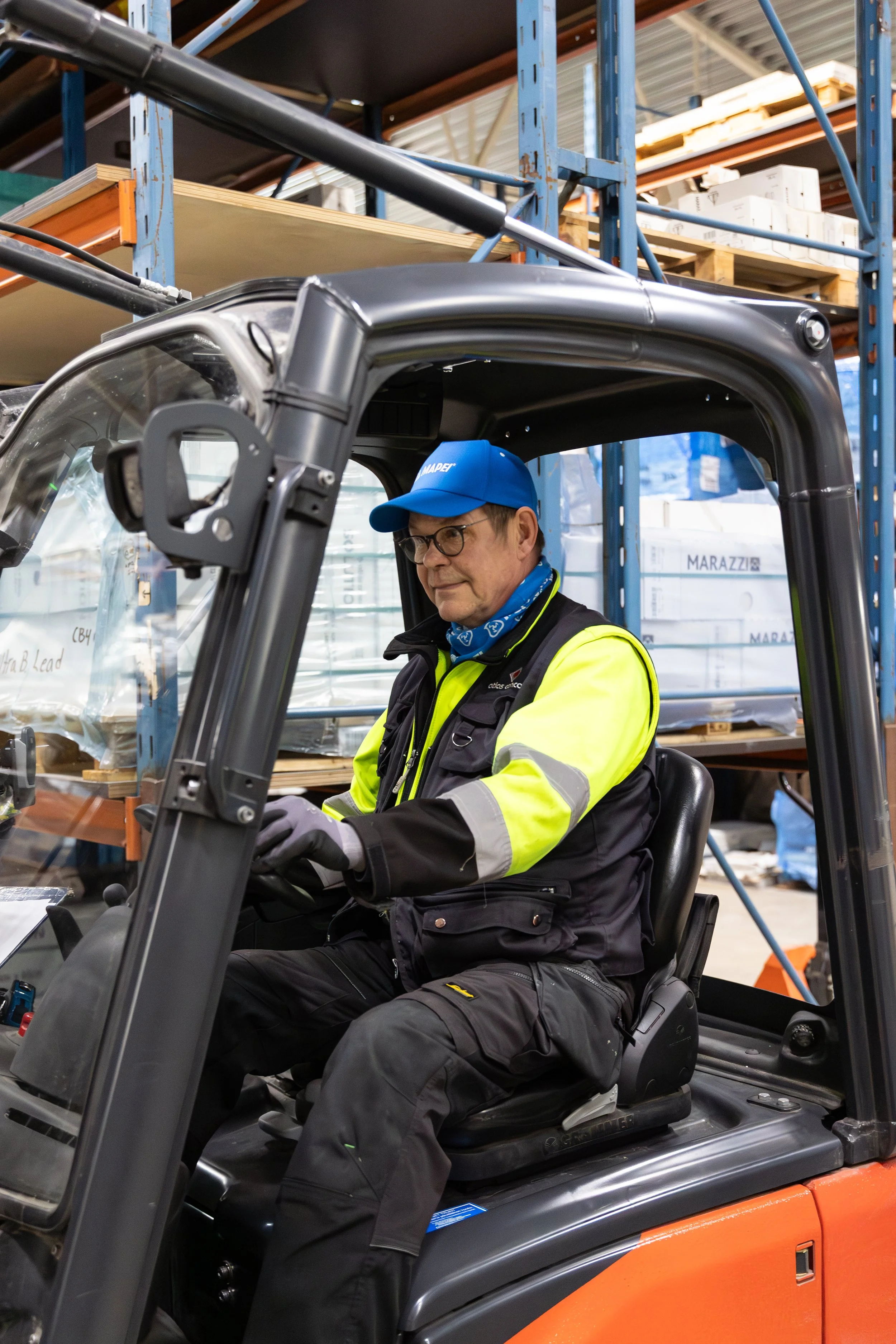 Mies kuljettamassa trukkia varastossa, taustalla hyllyjä ja varastotavaraa. Mies hymyilee.