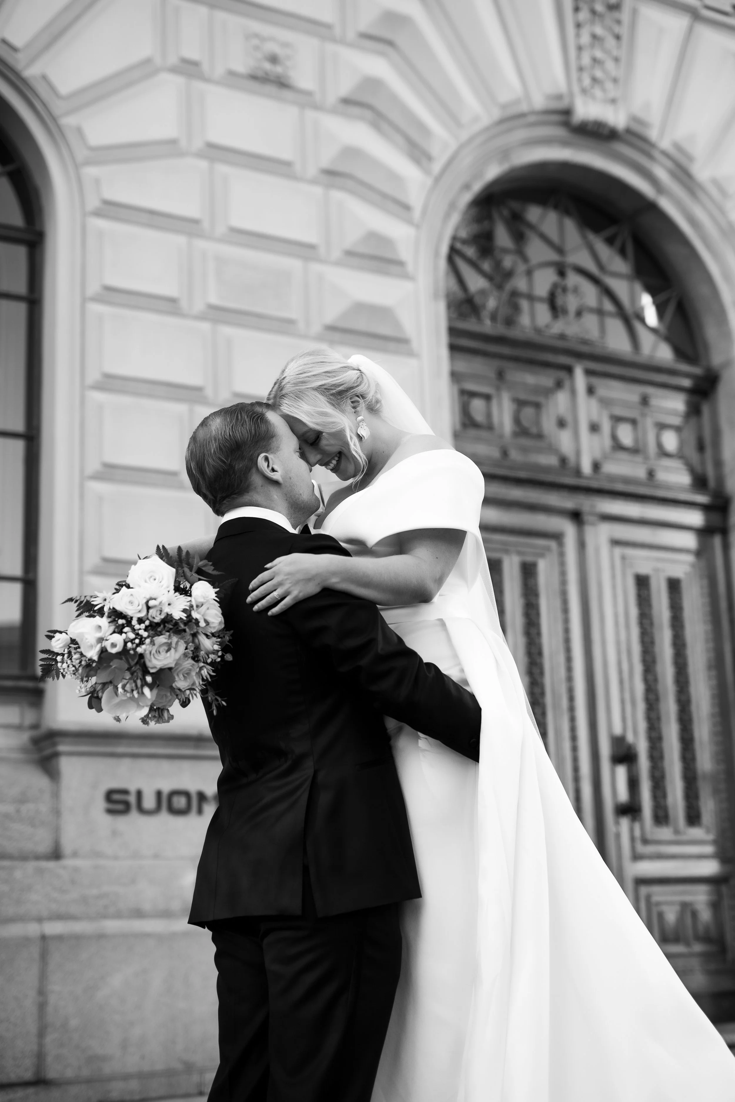 Hääpari syleilee toisiaan Suomen pankin edessä. Morsiamella on kaunis puku ja kukkakimppu. Morsian hymyilee leveästi.