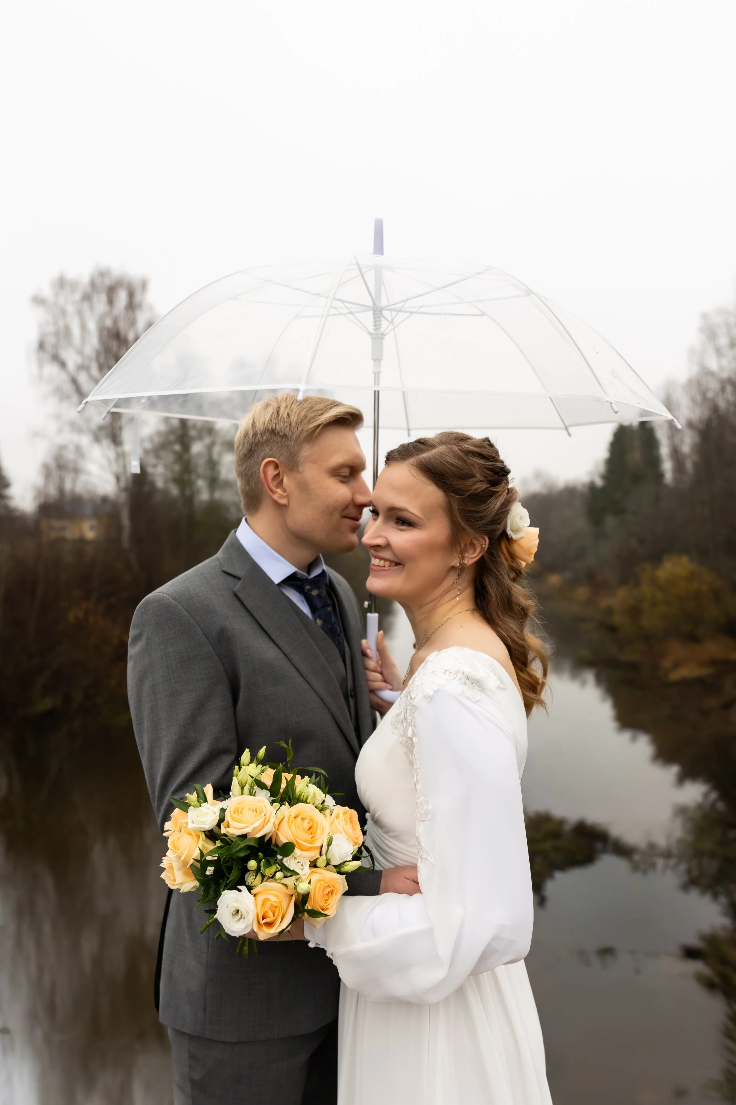 Hääpari seisoo sateenvarjon alla lähekkäin. Morsiamella on liehuva kaunis hääpuku ja sulhasella harmaa puku. Morsian pitelee kaunista kukkakimppua. Sää on hieman sateinen.