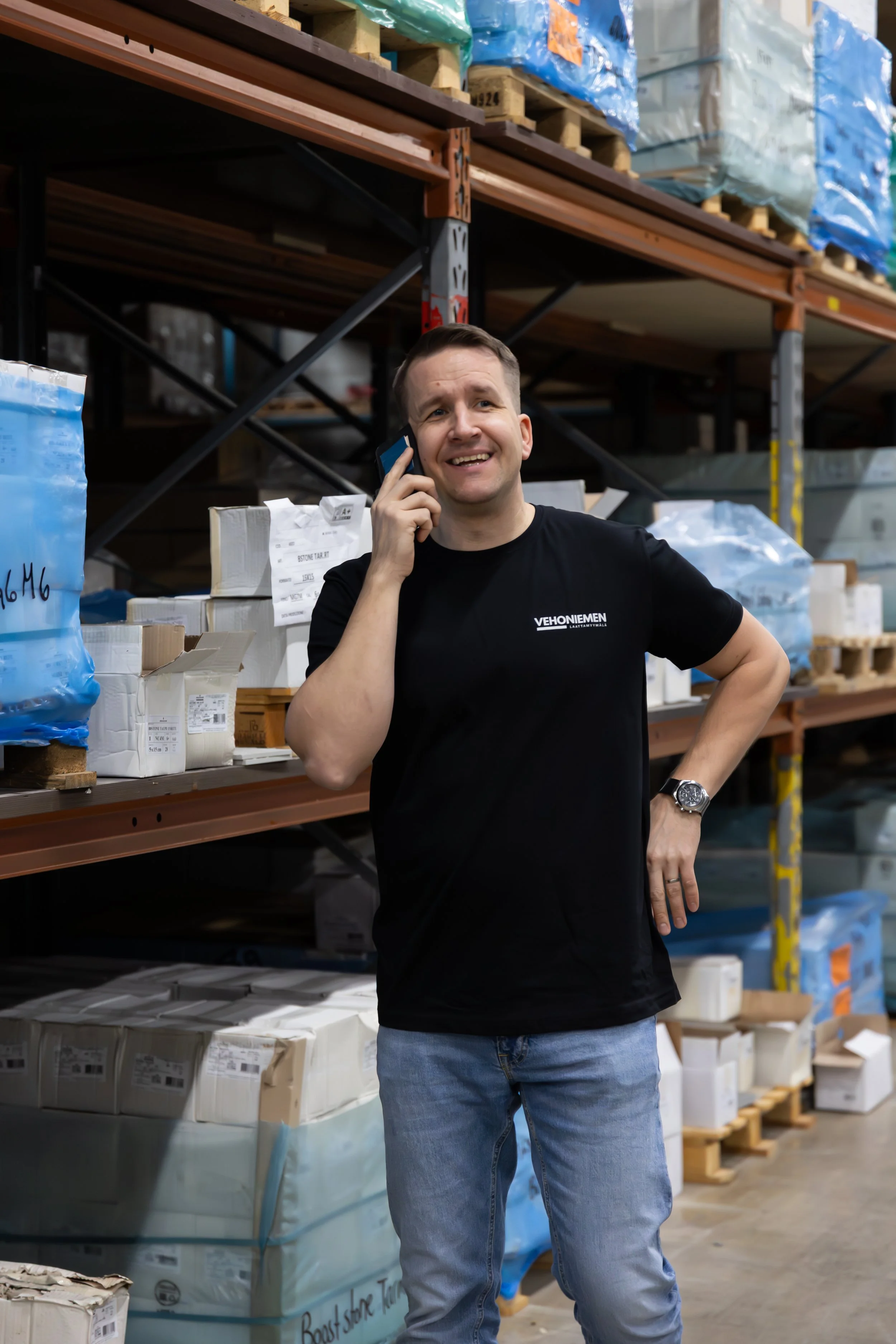 Mies puhelimessa varastossa, hymyilee, vaatteet, hyllyjä ja pakkauksia taustalla.