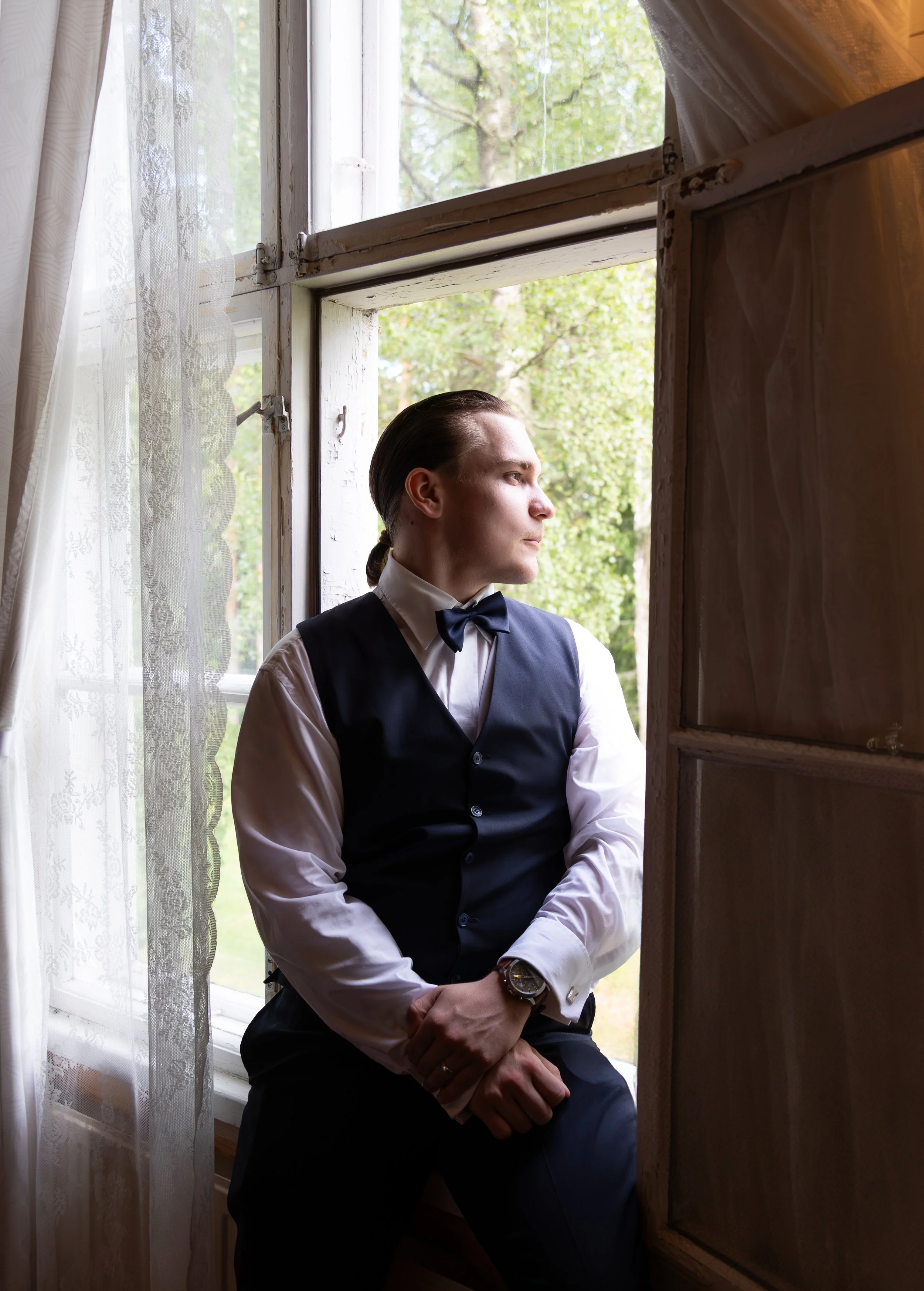 Mies istuu ikkunalaudalla, katsoo ulos ikkunasta, pukeutunut pukuun ja solmioon, taustalla vihreitä puita ja luonnonnäköala.