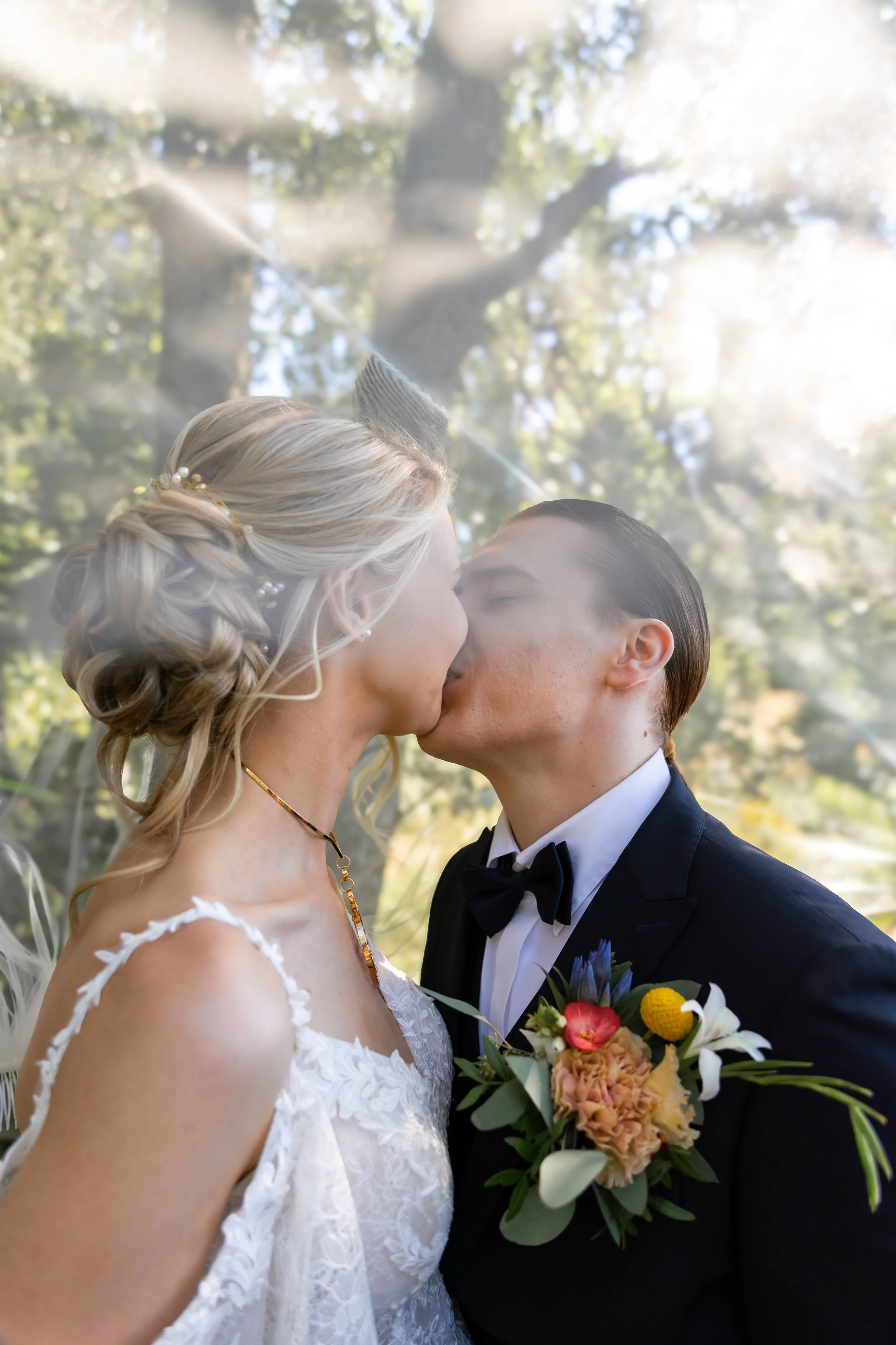 Hääpari suutelee toisiaan hunnun alla. Taustalla järvi ja kauniita puita. Morsiamella on kaunis hääpuku ja miehellä upea  musta puku.