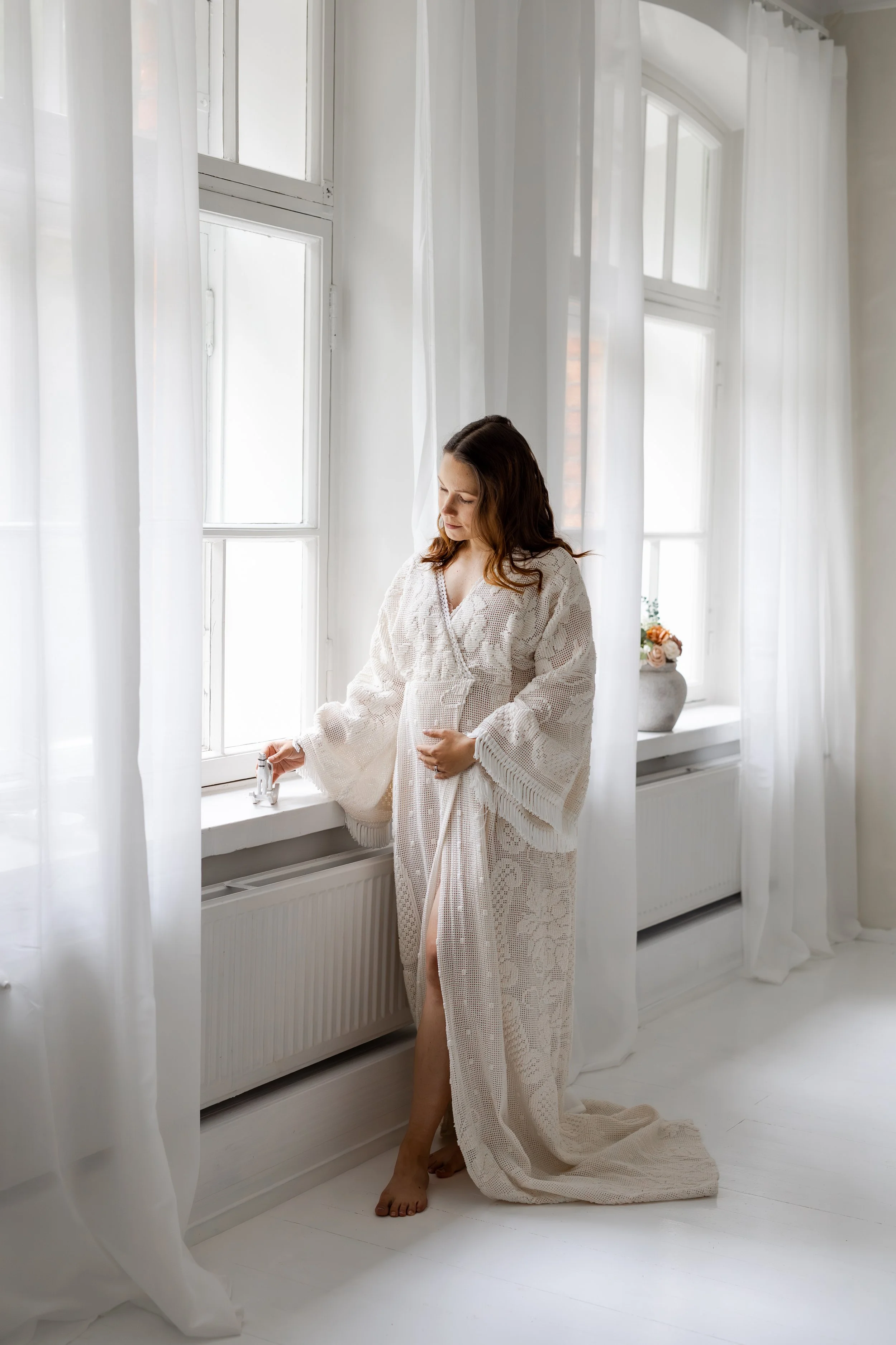 Raskaana oleva nainen seisoo ikkunan äärellä, pukeutuneena valkoiseen pitsipukuun. Nainen katselee lasten lelua.