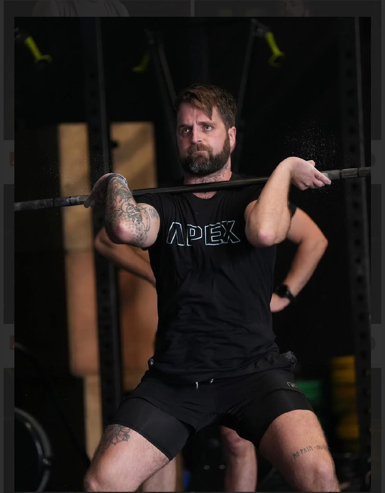 A man with a beard and tattoos wearing a black T-shirt and shorts is lifting a barbell in a gym, with another person standing behind him.