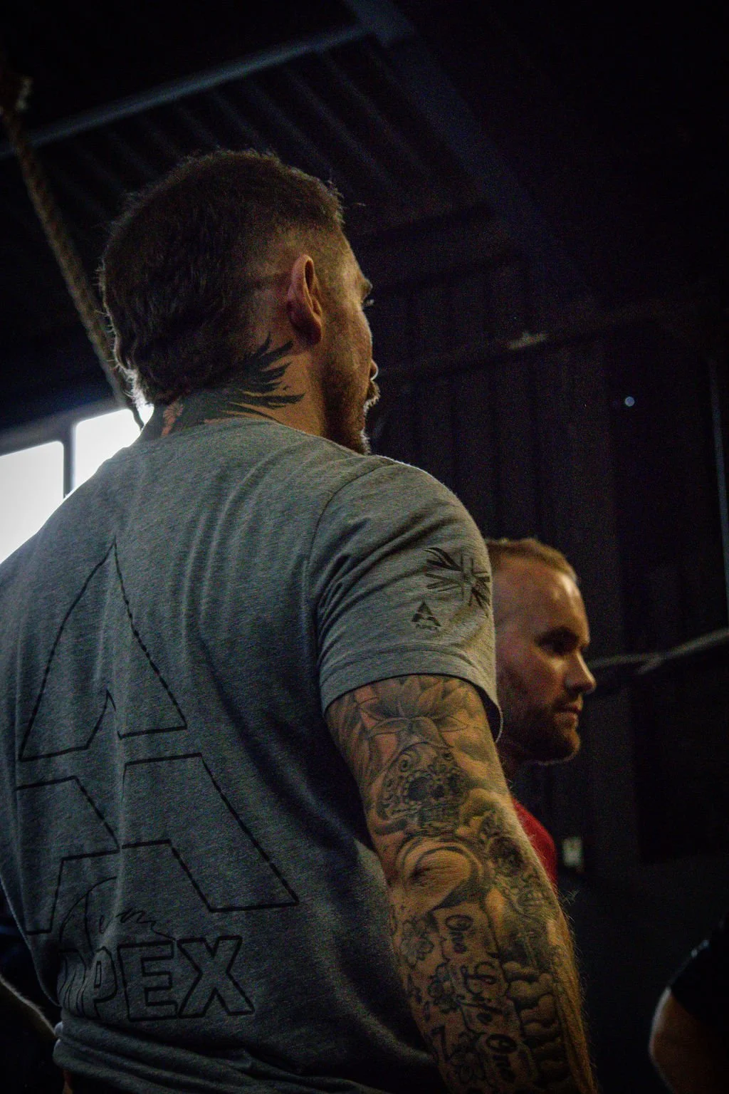 Man with short hair and tattoos on his arm, wearing a gray t-shirt, facing left, indoors with dark walls and roof structures visible.